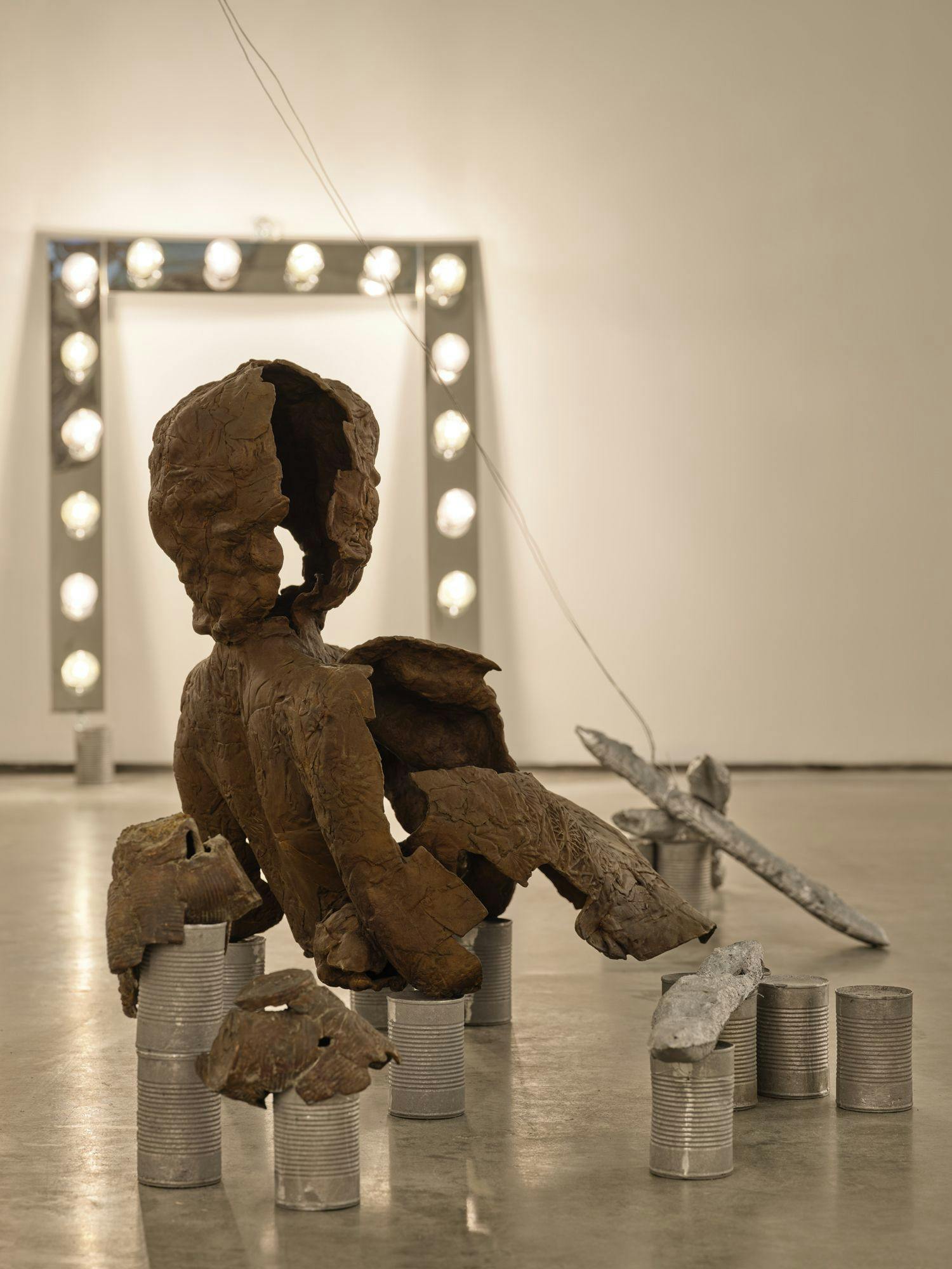 A bronze sculpture of a partial human figure sits on aluminum cans on a concrete floor. The sculpture's form appears to be precarious. Nearby are a vanity light, more aluminum cans, silver baguettes, and bronze cans that appear to be melting.