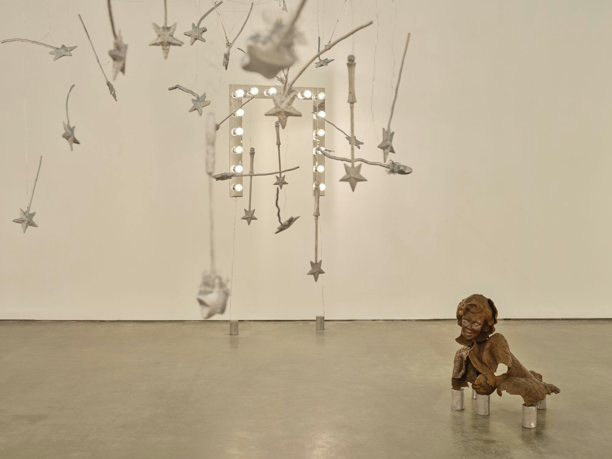 A wide view of several sculptures hanging from wire: magic wands, vanity lights and silver baguettes. A bronze sulpture of a partial human figure sits among aluminum cans and painted baguettes scattered across the floor.