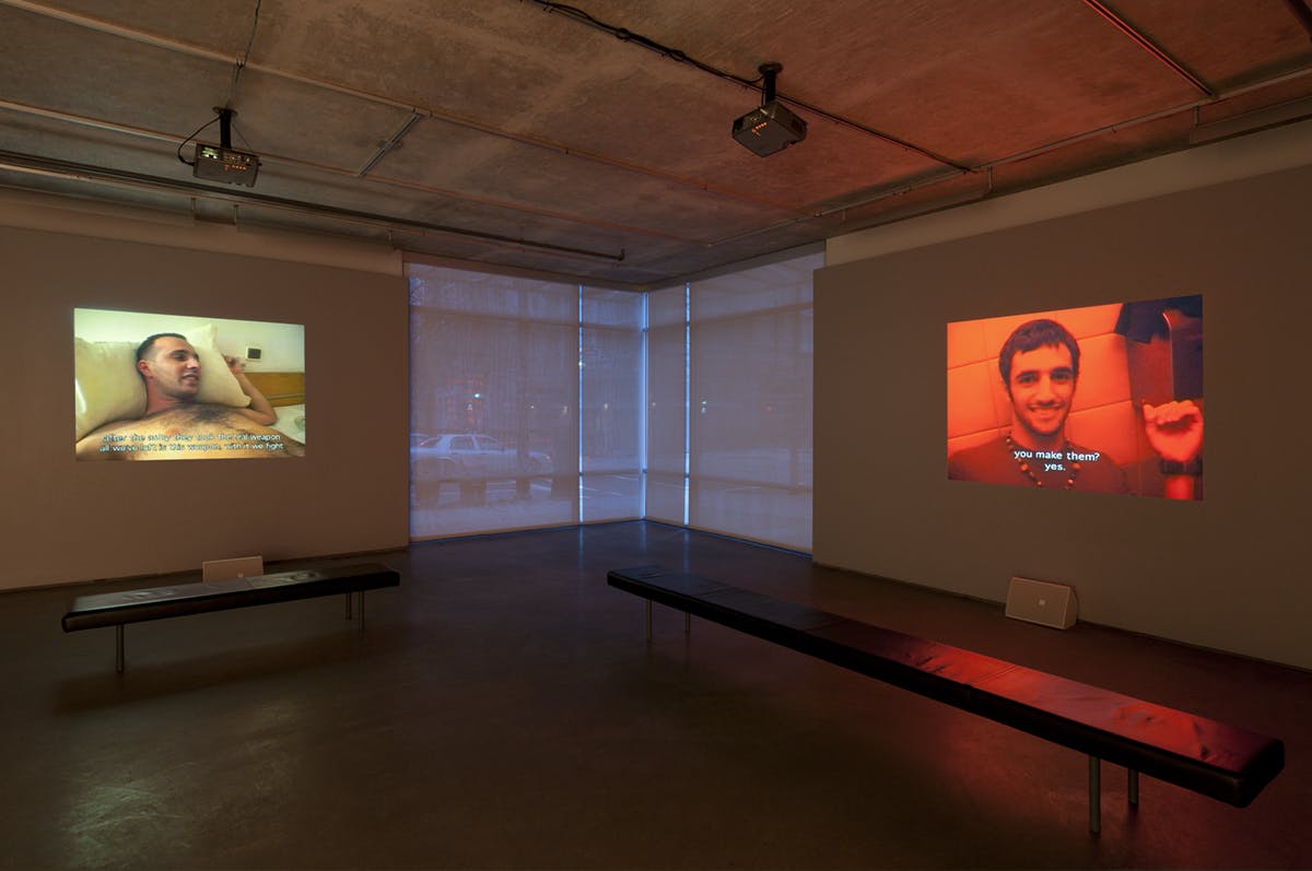 In a gallery space, two video works are projected on the walls. The video on the left shows the torso of a naked person lying on a bed, and another video shows a person smiling in a red-lit room.