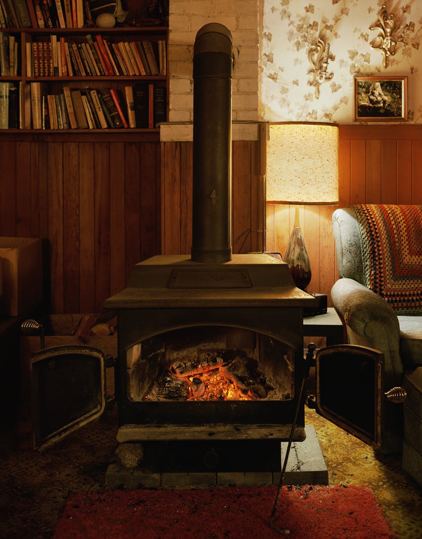 A photograph by Scott McFarland depicting the interior of a domestic space, which appears to be a living room with a wood burning stove, bookshelves, a couch and wood paneled wall. 