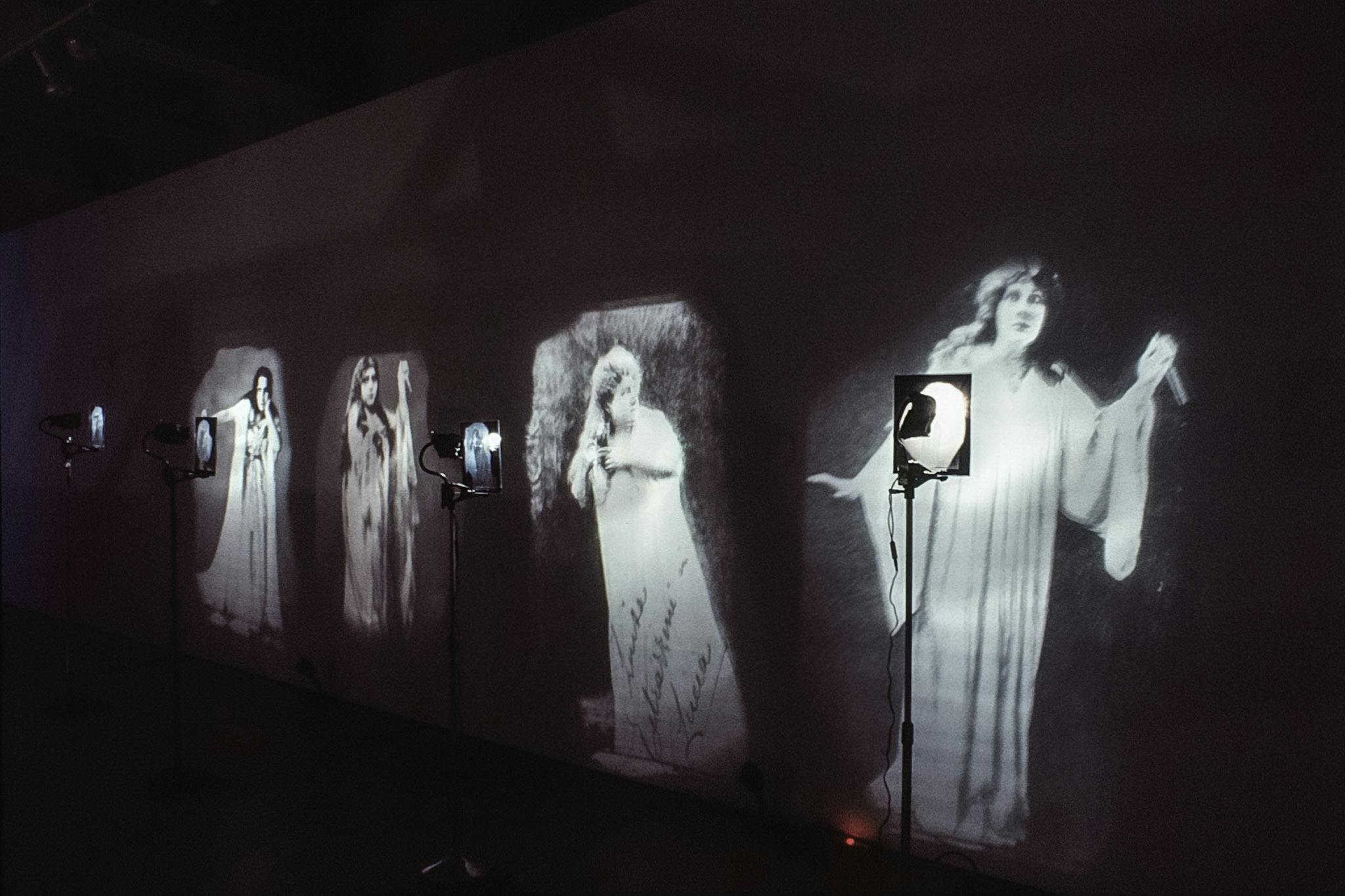 A series of four images are installed on a wall of a dimly-lit gallery. It is an enlarged  black and white photographs of women in white robes. A stage light is placed in front of each image of a woman.