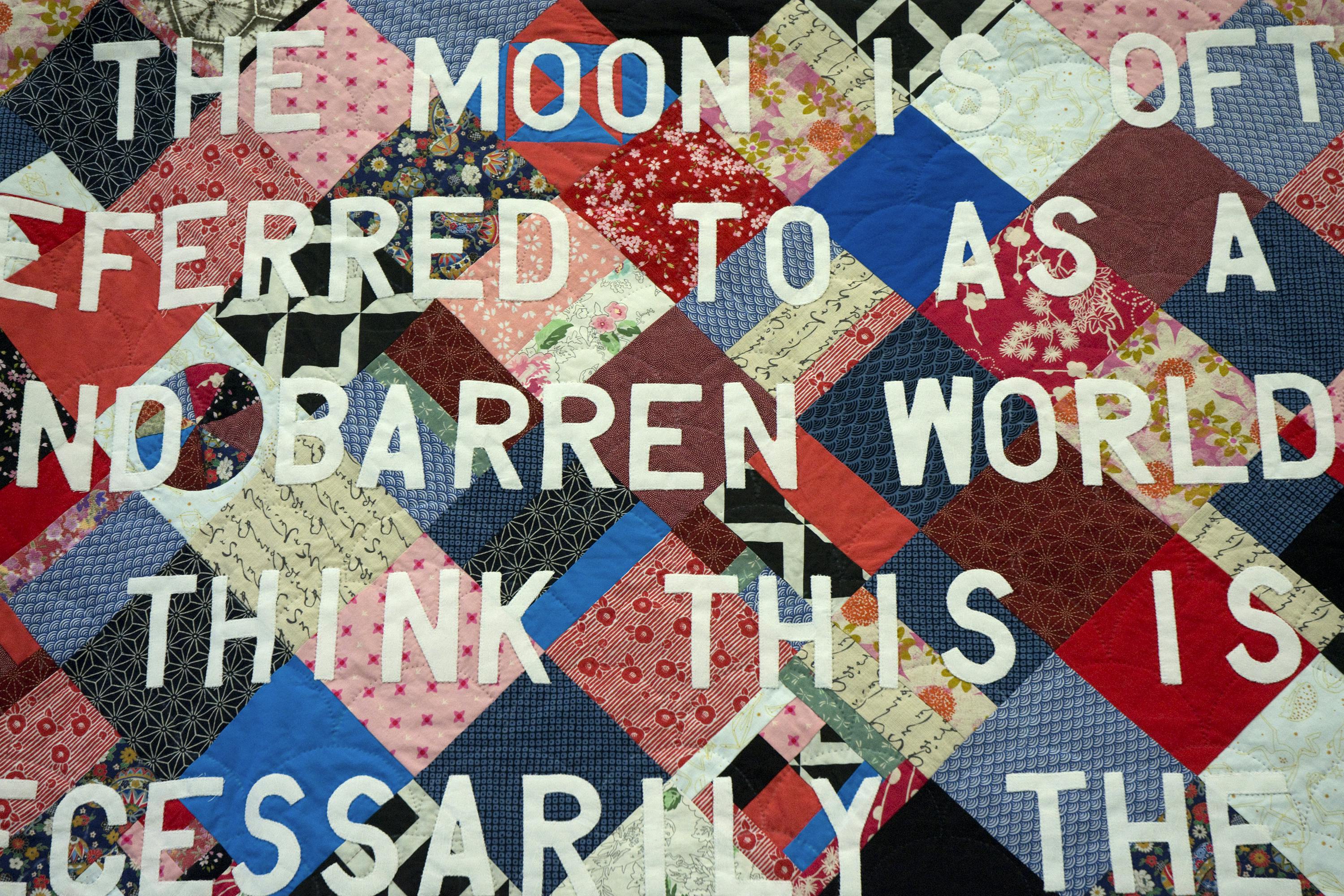 A close up view of a patchwork quilt made of fabric squares of varying patterns and colours. Quilted overtop are large white block letters. Only a fragment of a sentence is visible. 