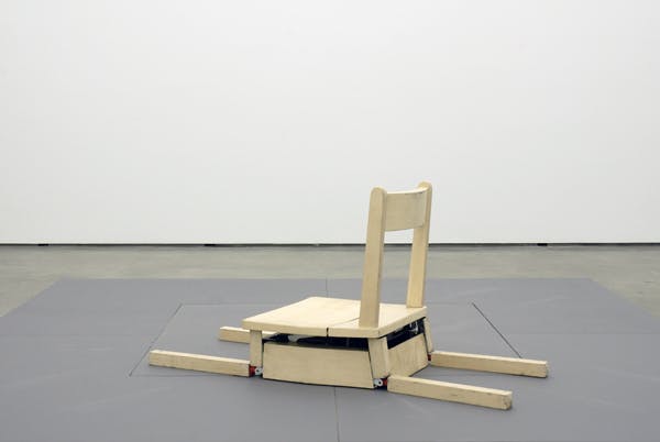 A small wooden chair is installed on the gallery floor. This chair is mechanicalized and moves automatically. In this photograph, the chair sits in the posture of a dog lying down.