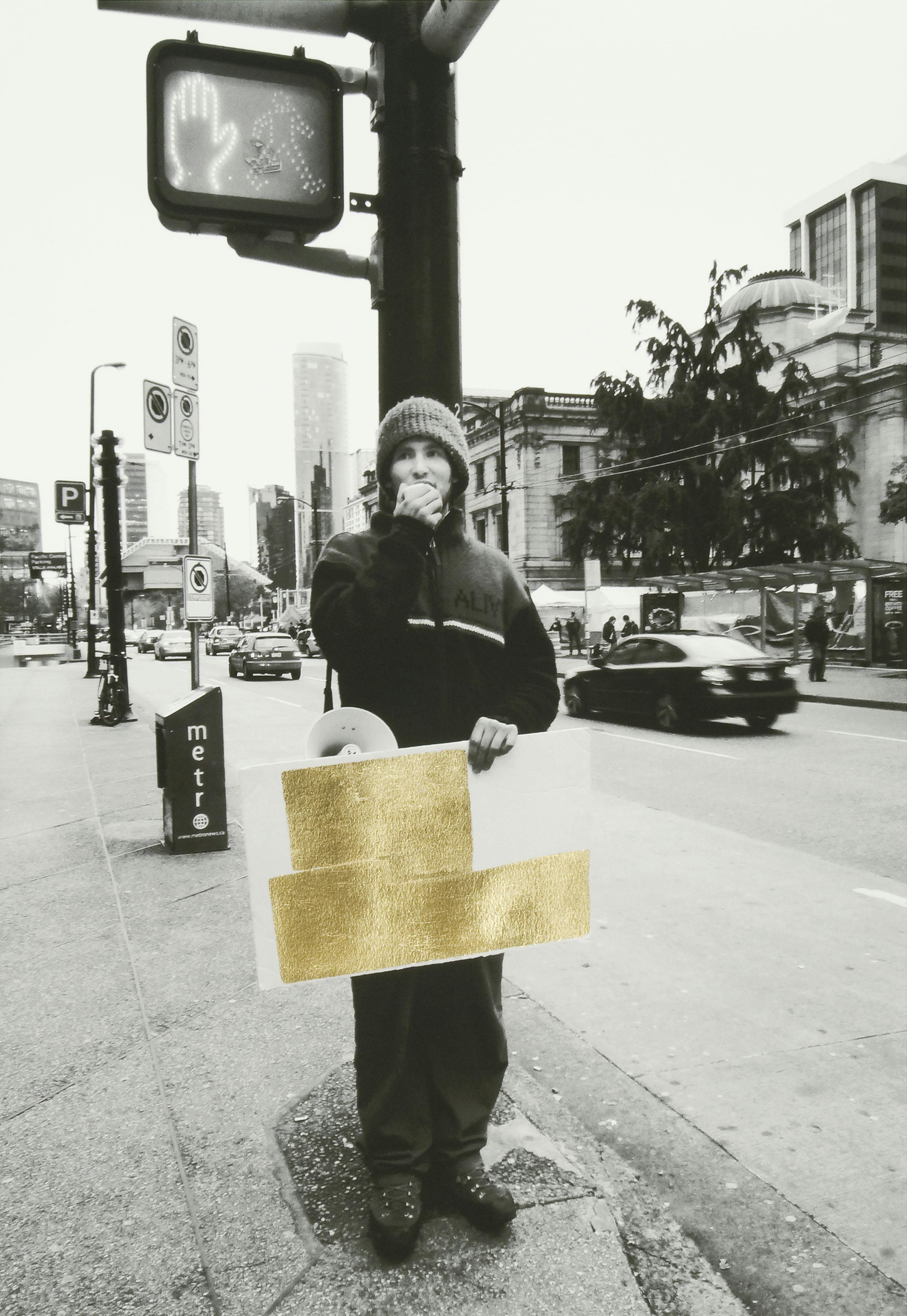 A black and white photograph of a person standing by a traffic light pole on a city street corner. They have a megaphone and are carrying a sign covered by a gold leaf intervention over the image.