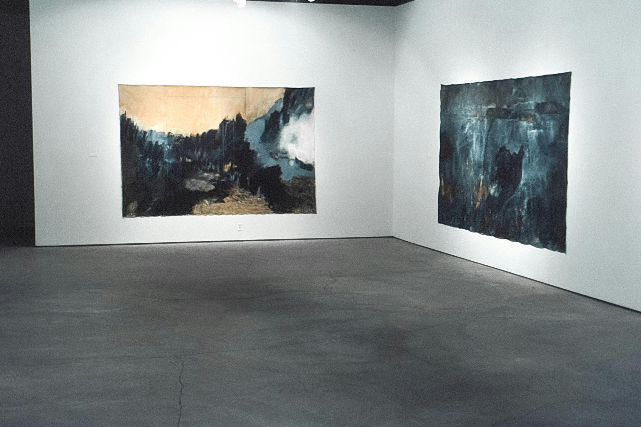 The corner of a gallery space, with one work on each wall. The works are large, dark paintings, with white, blue and yellow blended into them. They are on unstretched canvas, pinned to the walls. 