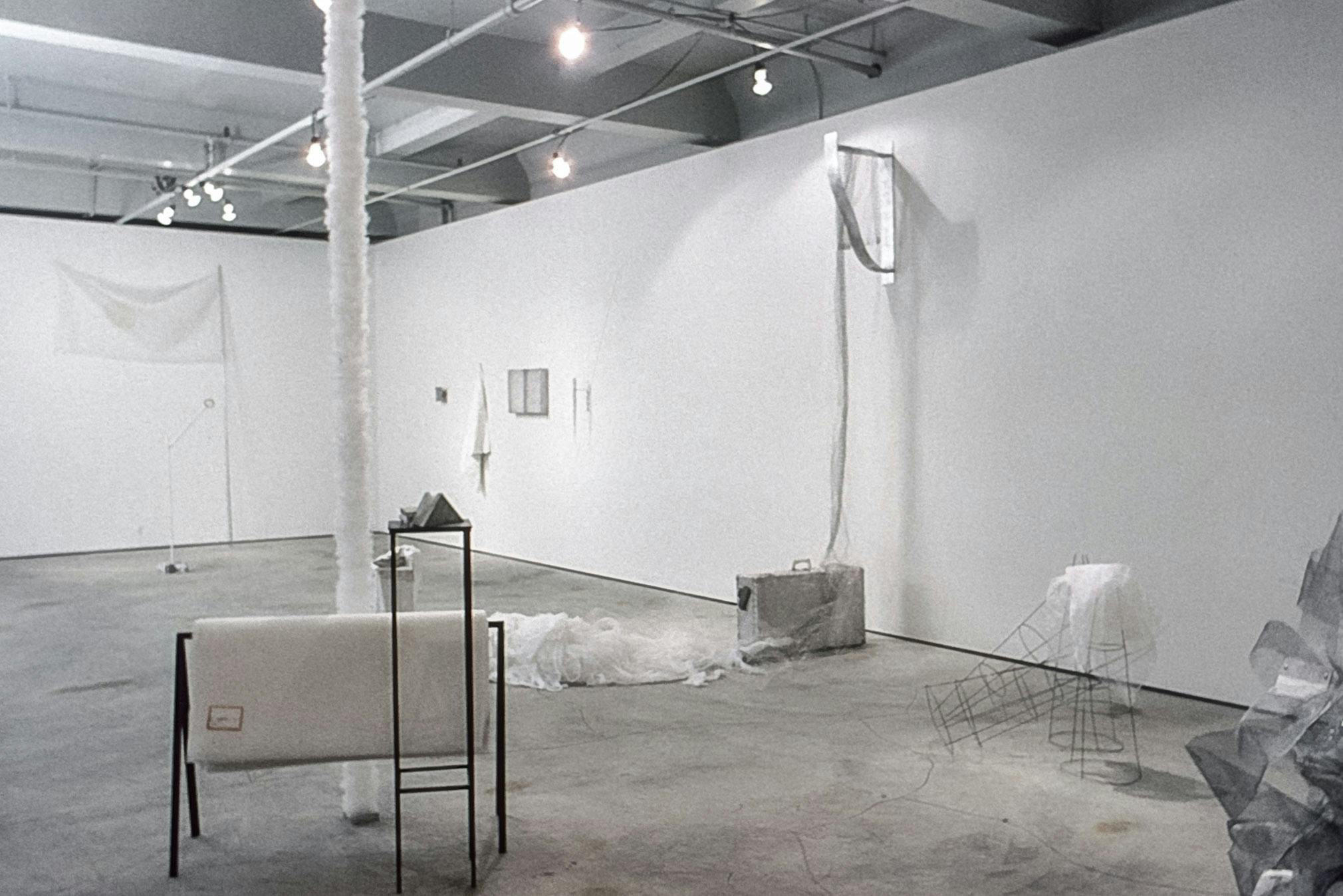A gallery with several artworks placed all around. The works include a white, textured tube shape hanging from the ceiling to the floor, and a mound of sheer white fabric on the floor. 