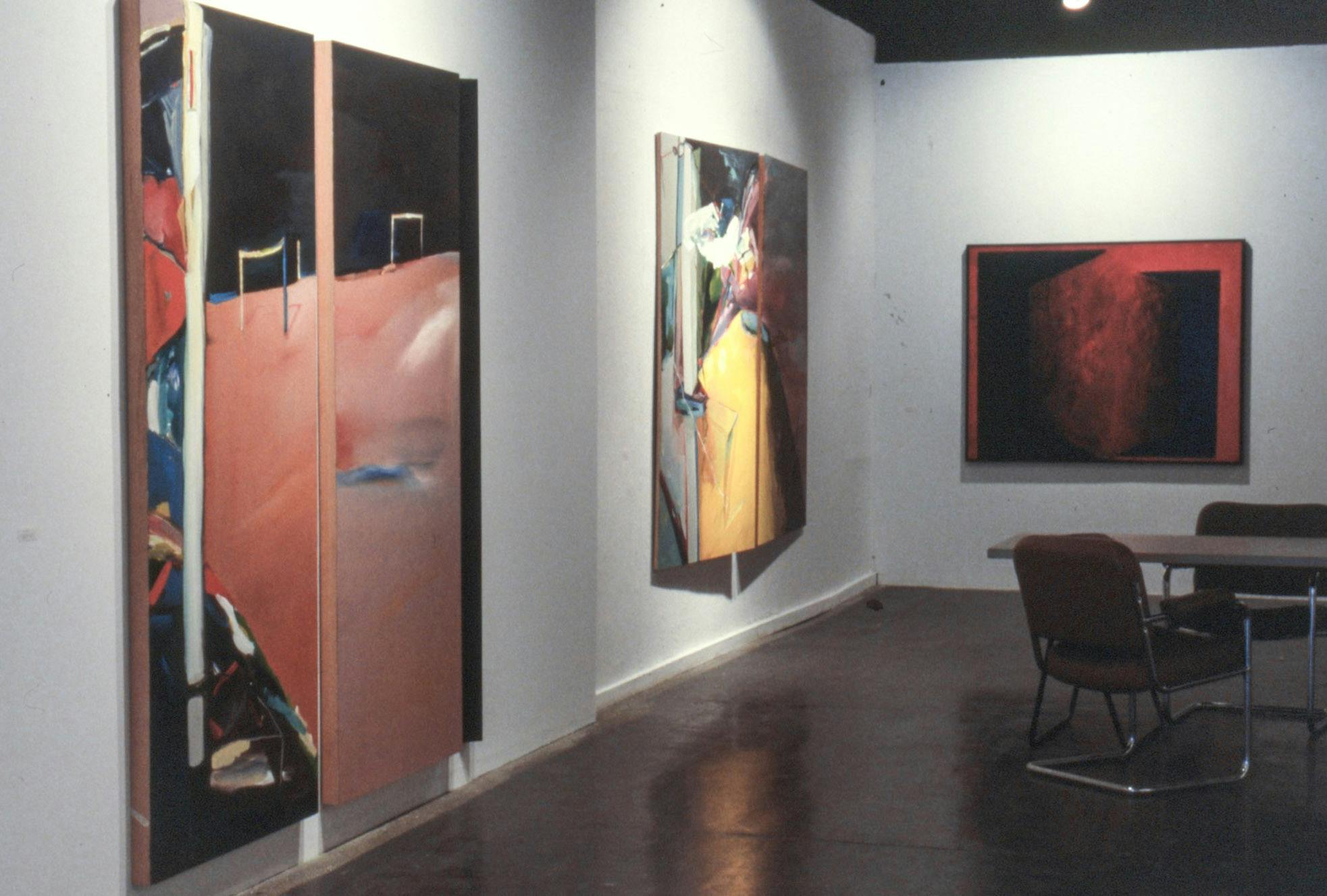 3 large paintings in a gallery. One is red and black in a frame. The other 2 paintings are bright and colourful, composed of 3 different canvases. On the floor, there is a table and 2 chairs visible.