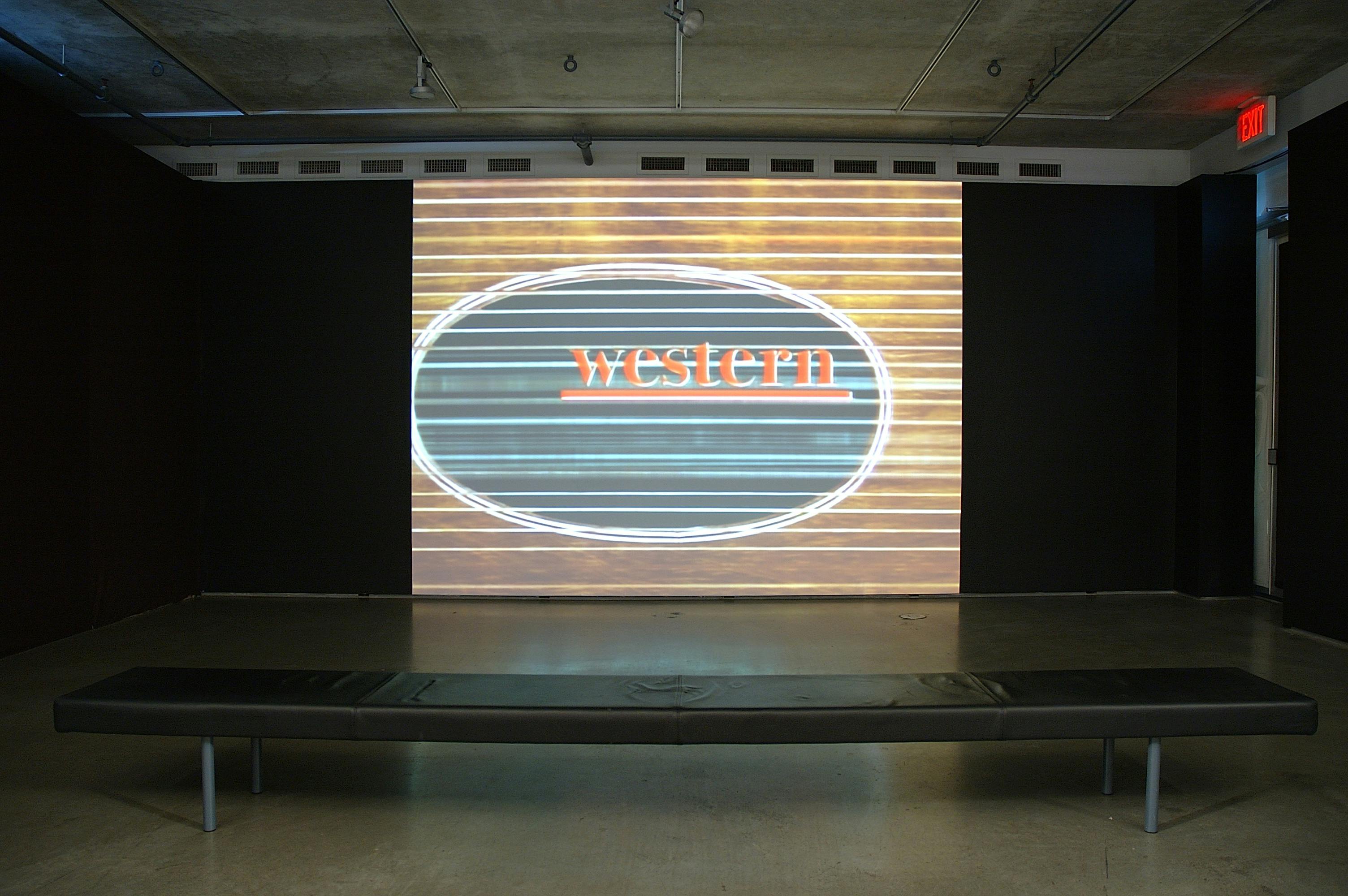 An installation image of a video projected on a wall in a dimly lighted gallery. The video shows a word "western."