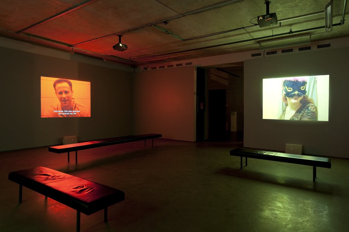 Two video works are projected on gallery walls. The video on the left shows a person saying, “Because the sex started to control my life.” Another video shows a person smiling under a black cat mask.