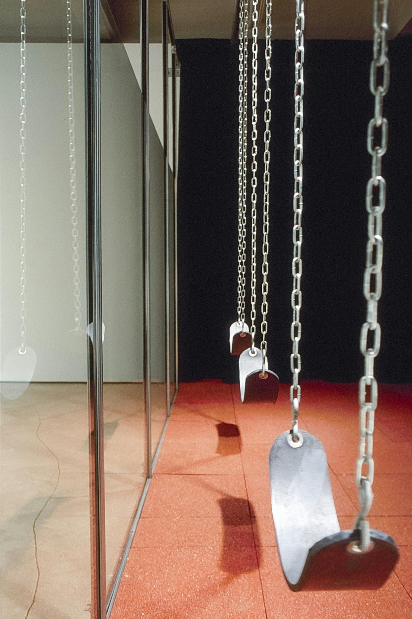 An installation view of three swings in the art gallery space. The black rubbered seats with silver chains are hung from the ceiling. The swings are placed right next to the large glass window.