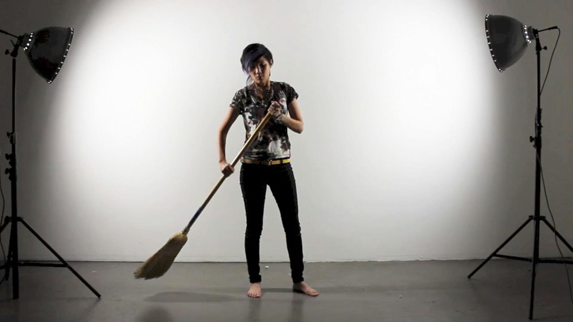 Still image from a video. A person wearing a grey shirt and a pair of black pants stands on a concrete floor between spotlights, sweeping with a long broom. 