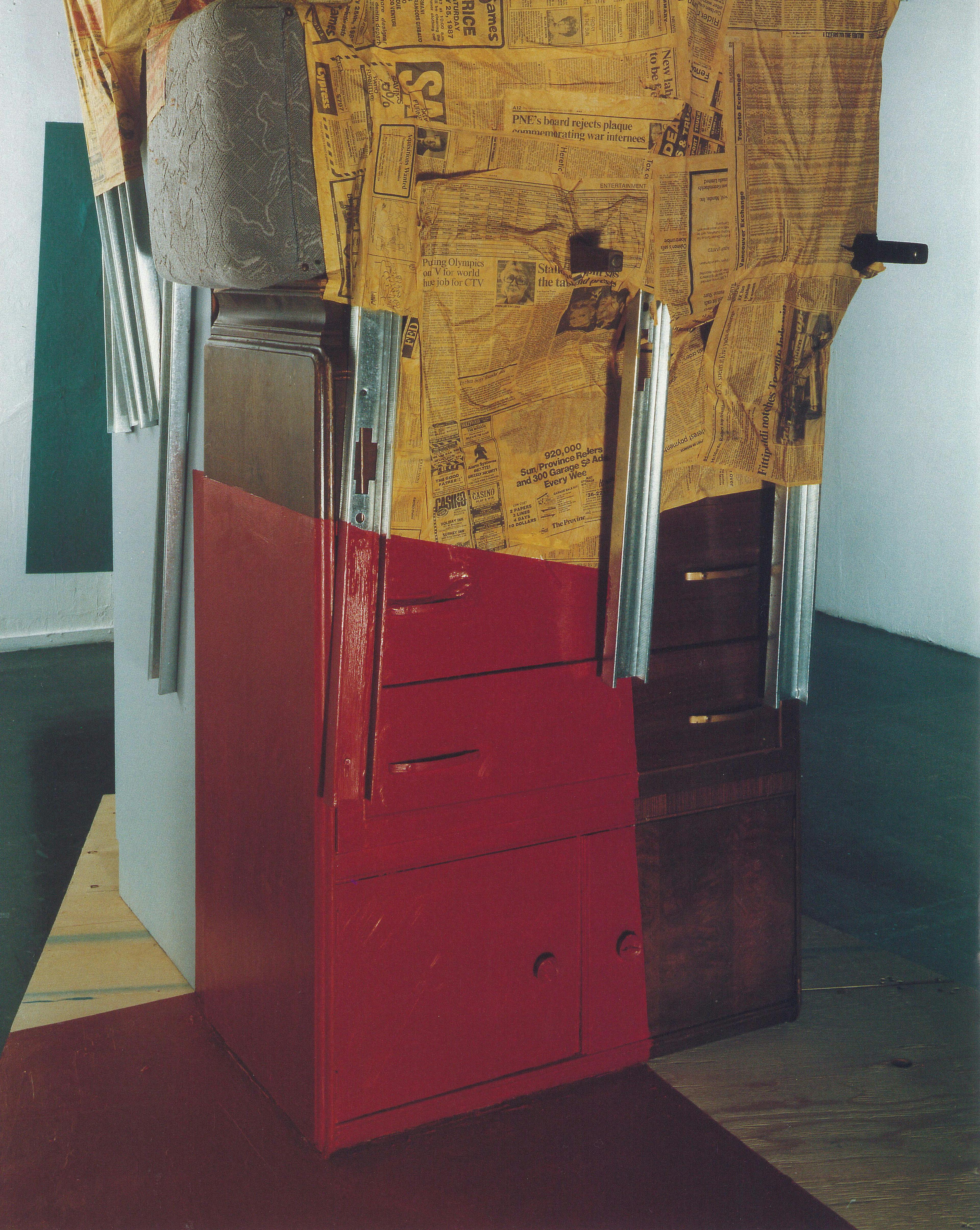 This is a close-up view of one of Jessica Stockholder’s sculptures installed in a gallery. A red-painted drawer sits on the floor. The top half of the furniture is covered by old newspapers. 