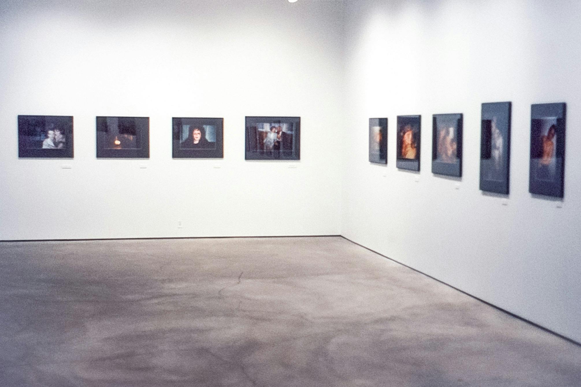 The corner of a gallery space. Mounted on the white walls are photo works in black frames . The photos are of people in dark rooms. Some are embracing, some are dressed, and some are half-dressed.