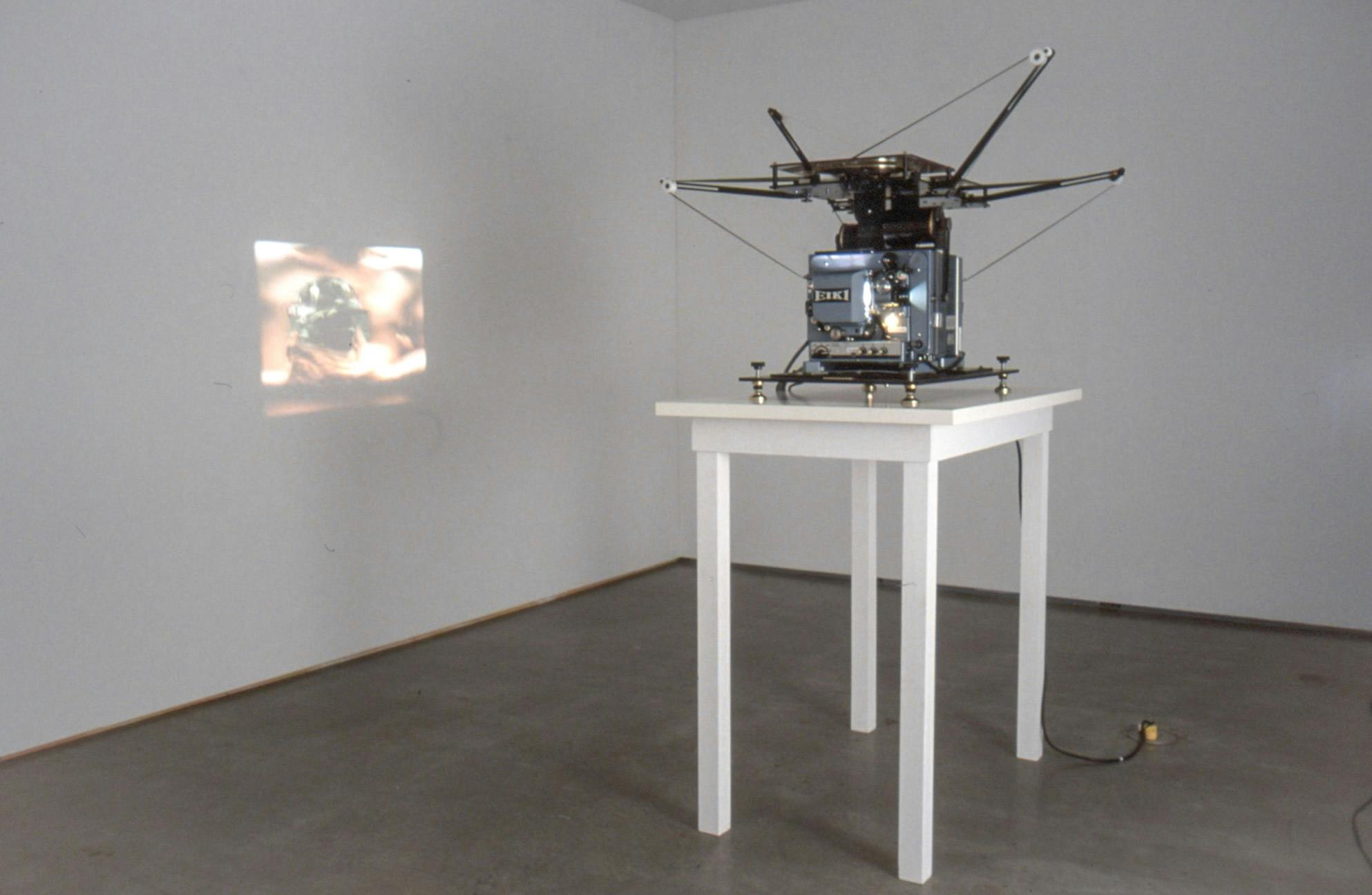 A film projector projects a moving image on a white gallery wall. The film shows a torso of a naked person holding a video camera to a person who was filming. 