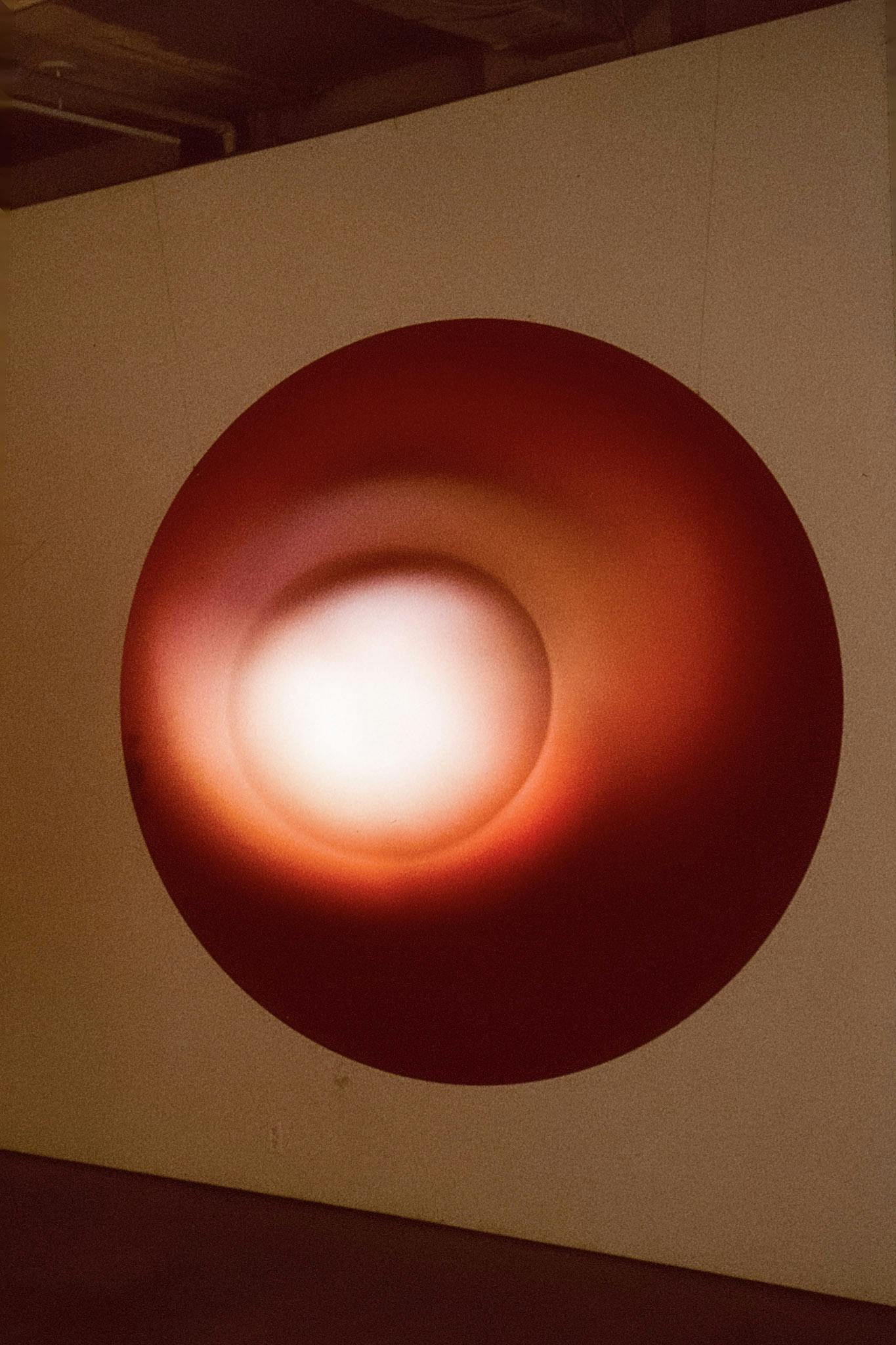 A large circular-shaped red sculpture is installed in the gallery. The sculpture is thin and completely attached to the wall. There is a circle of white light glowing in the middle of the sculpture.  