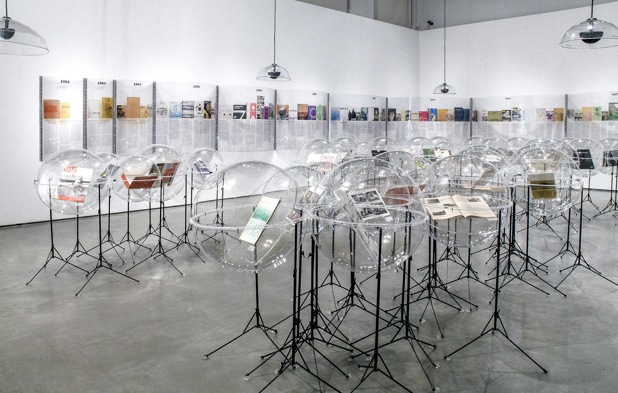 Many sculptures are installed in a gallery. They are magazines placed inside transparent rounder-shaped plastic cases. Those cases are supported by black metal legs similar to those of music stands. 