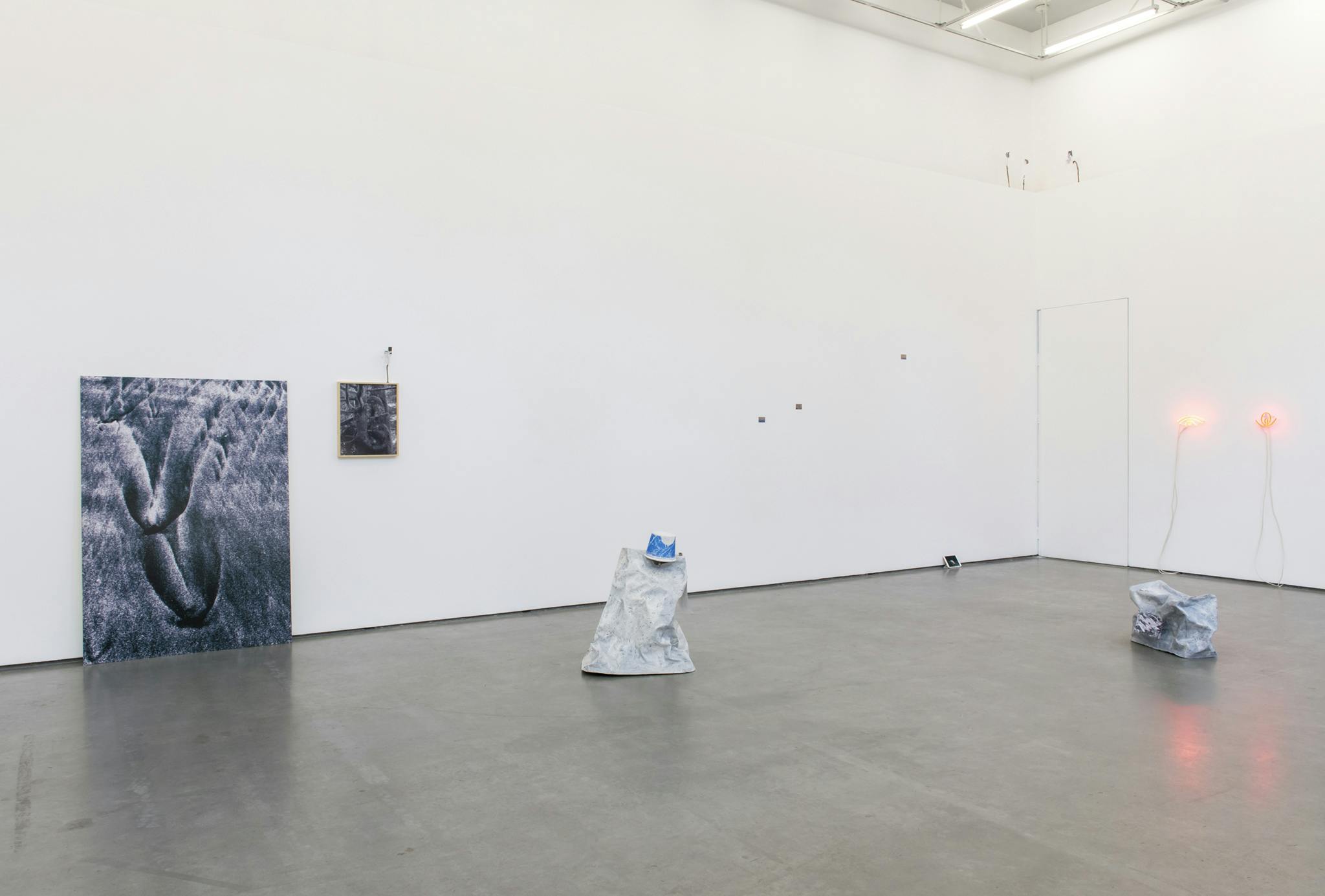 Installation image of the group show The Blue Hour. There are various works that range from sculptural pieces sitting on the floor, neon light fixtures installed on the wall, and photographic works. 