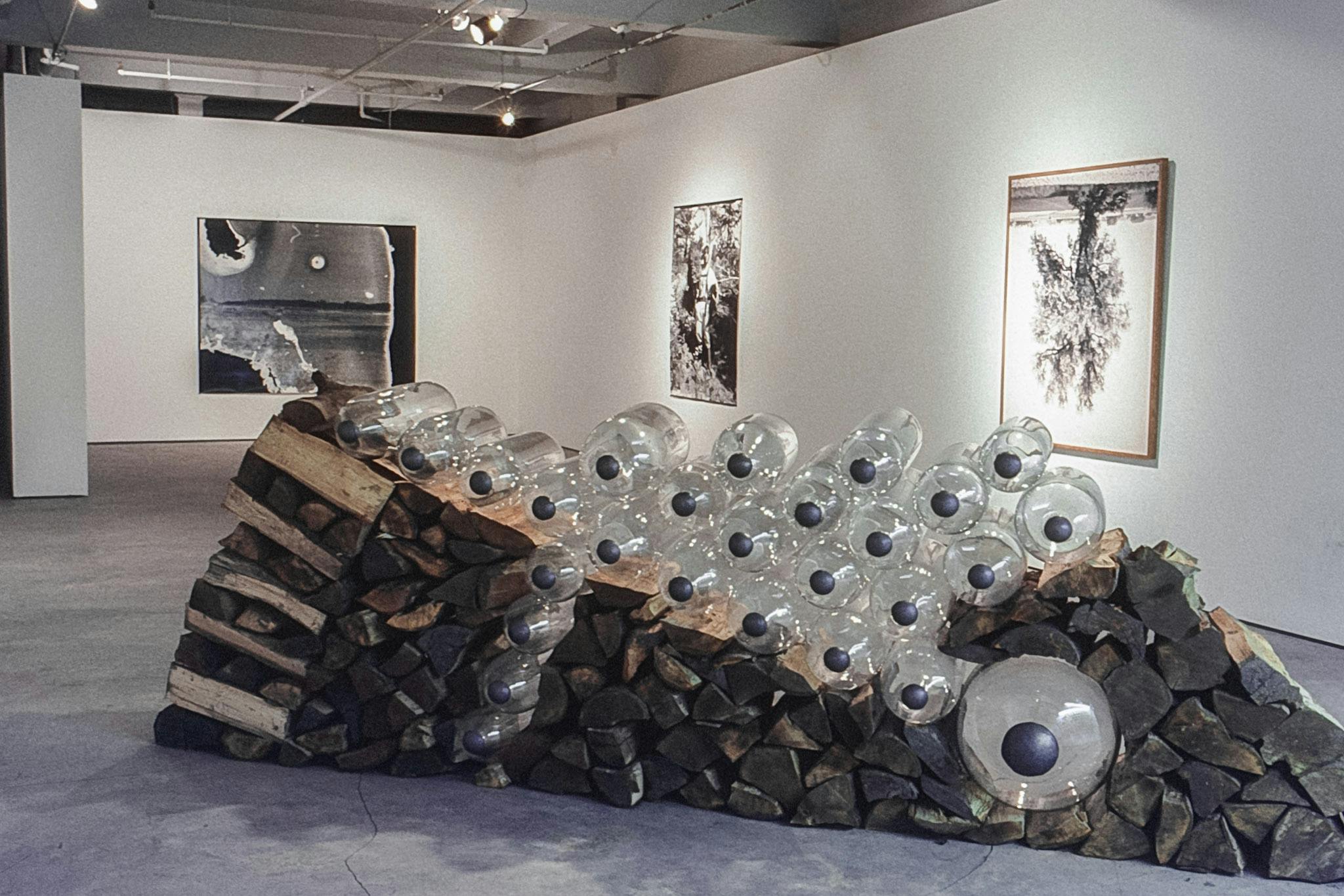 A closeup image of a large artwork. The work is made of large glass cylinders of different sizes and firewood. In the background, three large black and white photos can be seen on the walls.