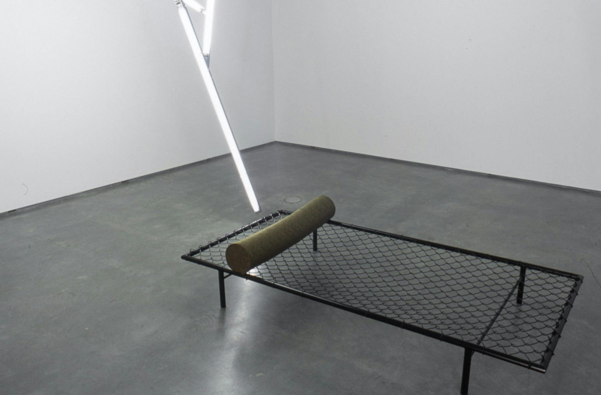 A black bed frame is placed on the gallery floor. A cylindrical form is placed at the edge of the bed frame. A sculpture made of tube light bulbs is partially visible behind the bed frame. 