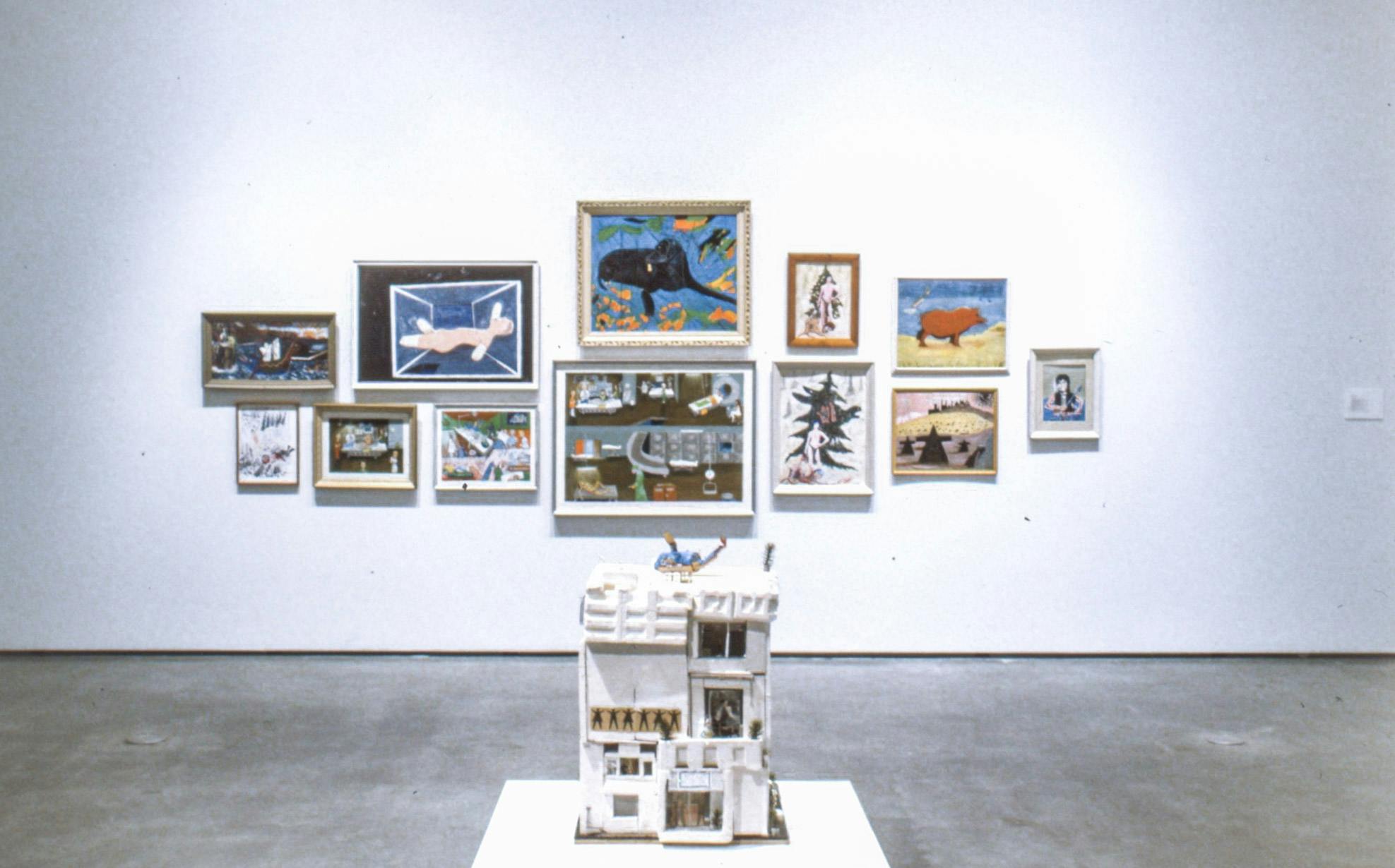 Twelve framed paintings are mounted on the gallery wall behind a sculpture. The sculpture depicts a white apartment building, on which a blue helicopter is landing.  