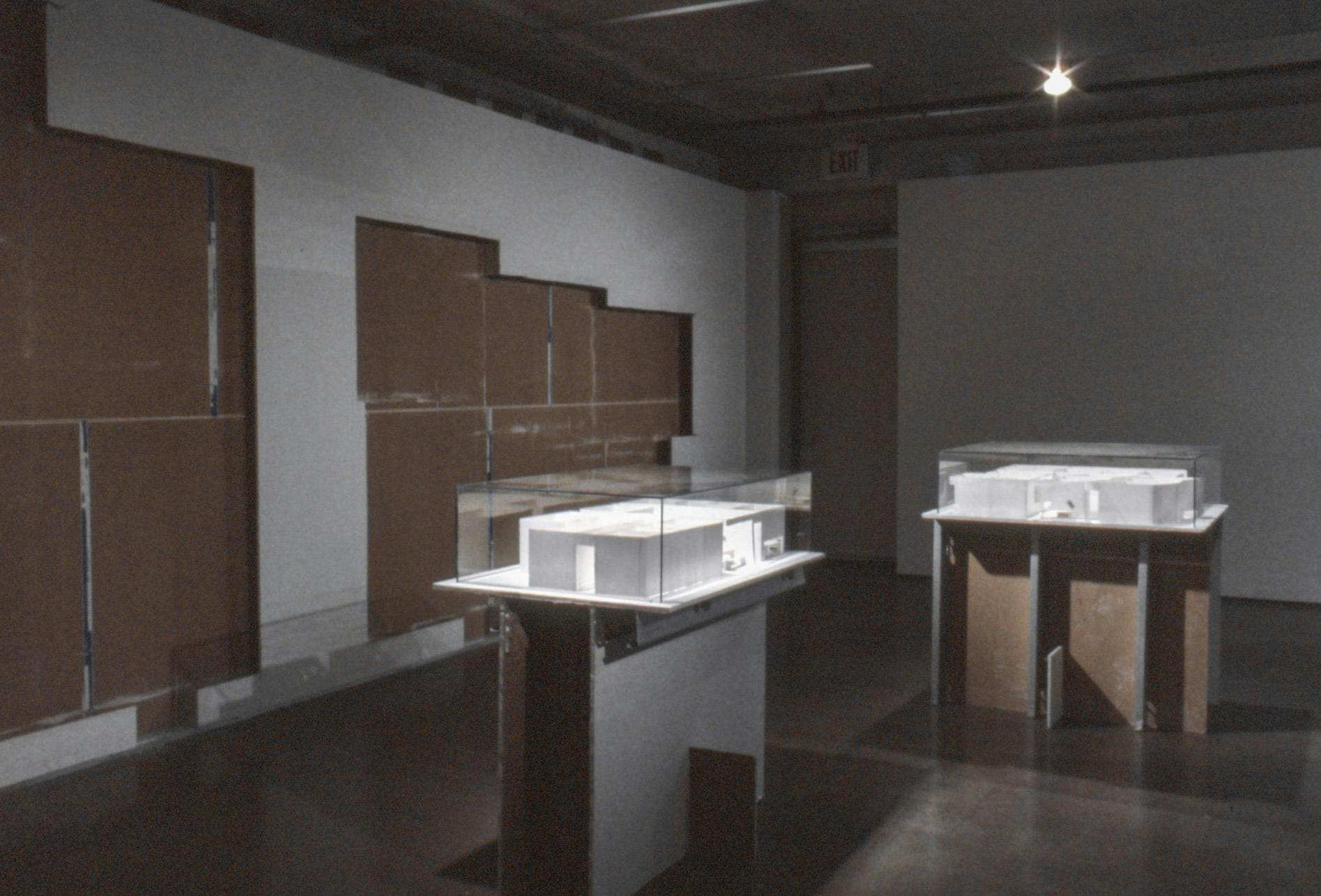 A set of white sculptures in display boxes are placed in a gallery space. White tiles on one of the gallery walls were removed, making the brown internal part of the wall visible. 