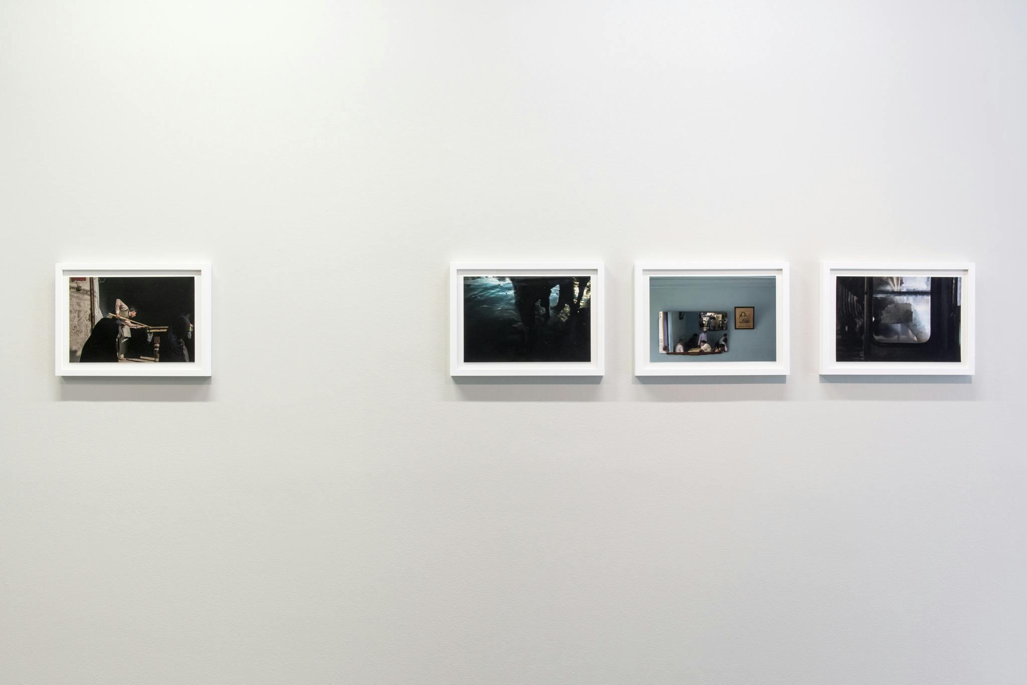 Four framed photographs hang horizontally on the wall of the gallery. Three photographs are grouped together on the right, the fourth sits alone on the left. The frames of the photographs are white.