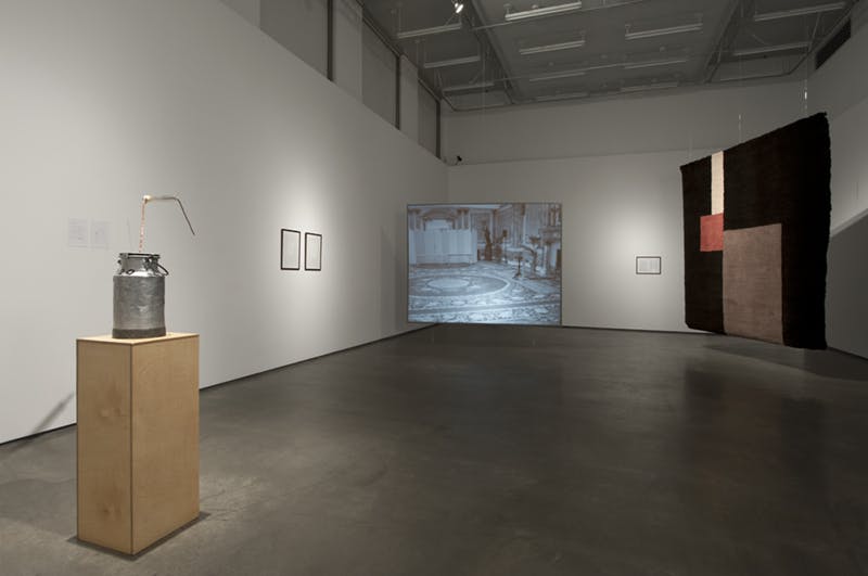 Sarah Browne's artworks installed in a gallery. A large, hand-knotted carpet hangs from the ceiling next to a film projected on a screen. A sculpture resembling a milk can sits atop a wooden plinth.