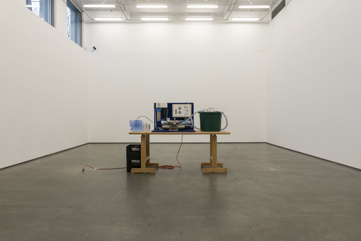 A series of objects appear on a large wooden table in the middle of a gallery space. From left to right, the table includes a water jug, stacks of drinking glasses, a machine, and a large green bucket.