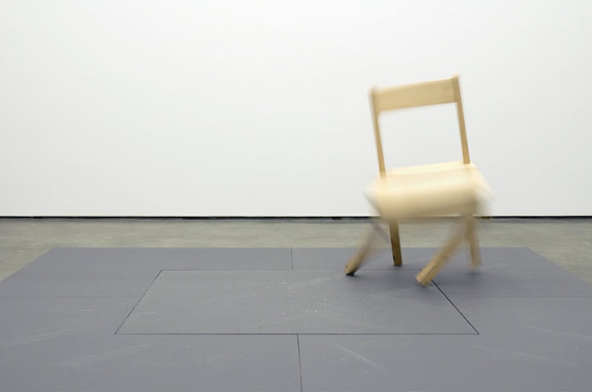 A small wooden chair sits on the gallery floor. This chair is mechanicalized and moves automatically. In this photograph, the front two legs of the chair are quickly shifting to its right. 