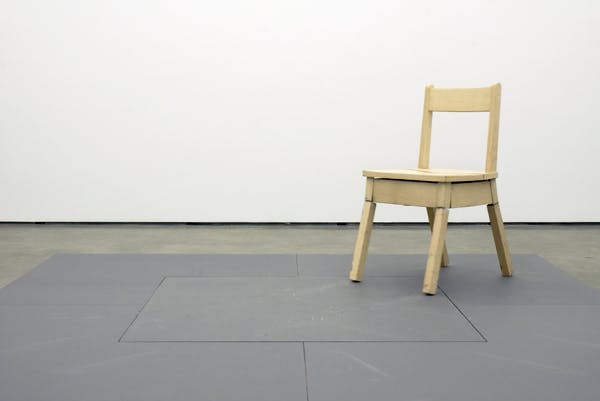 A small wooden chair sits on the gallery floor. This chair is mechanicalized and moves automatically. In this photograph, the chair is staying still, or it is about to move its legs.