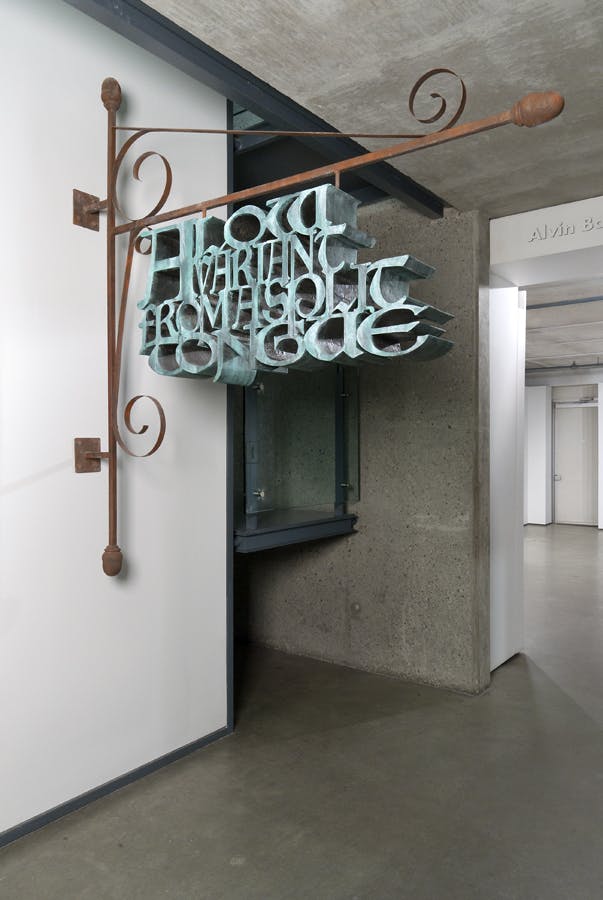 A large-scale iron sign mounted on a gallery wall. The sign reads A Low Variant From A Split Tongue in turquoise painted iron. The phrase hangs from an L shaped bracket that is mounted to the wall.