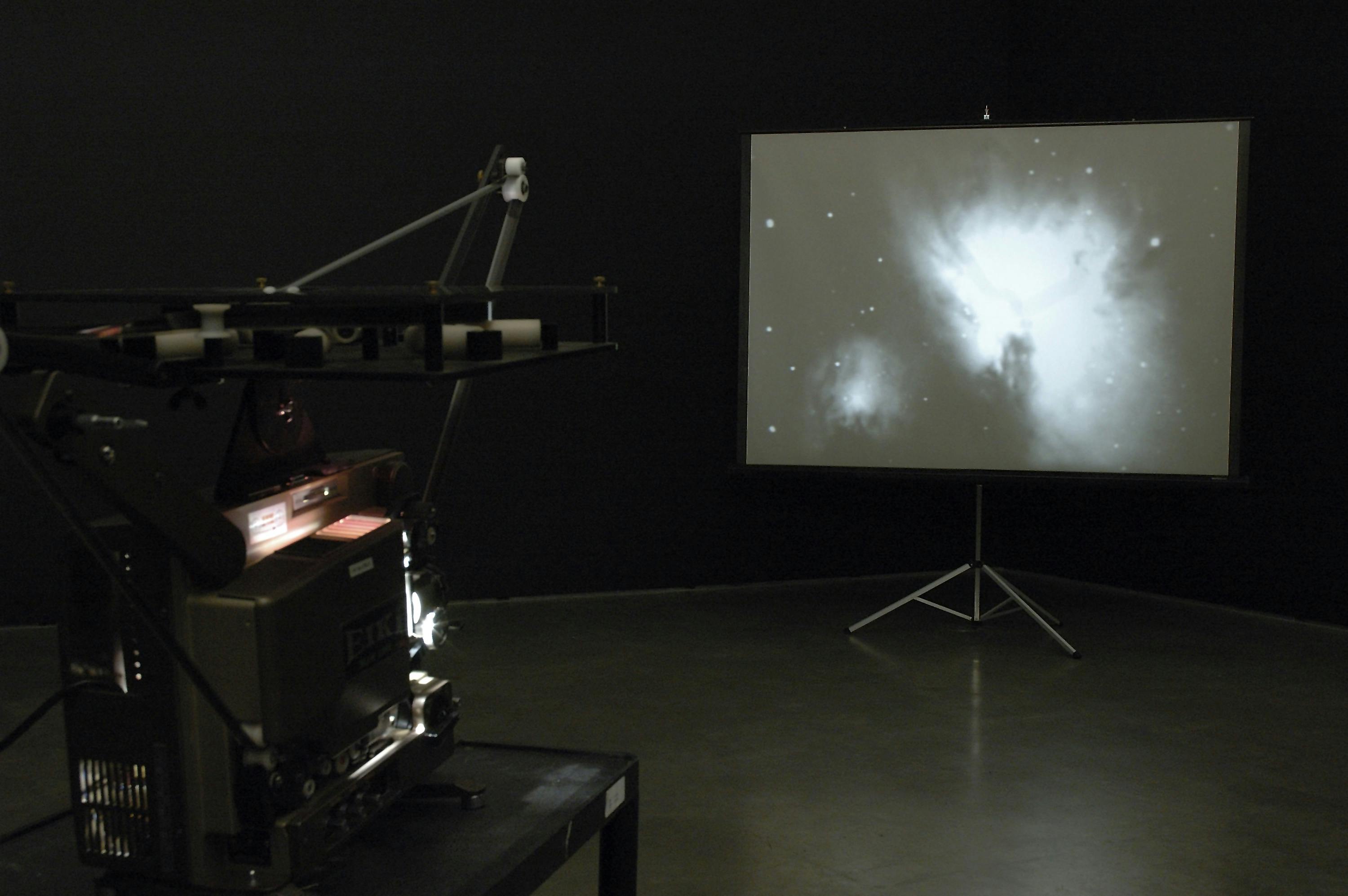 A film projector is installed in a darkened gallery space. It projects an image of white-blue stars and a nebula on a projector placed in the corner of the gallery. 