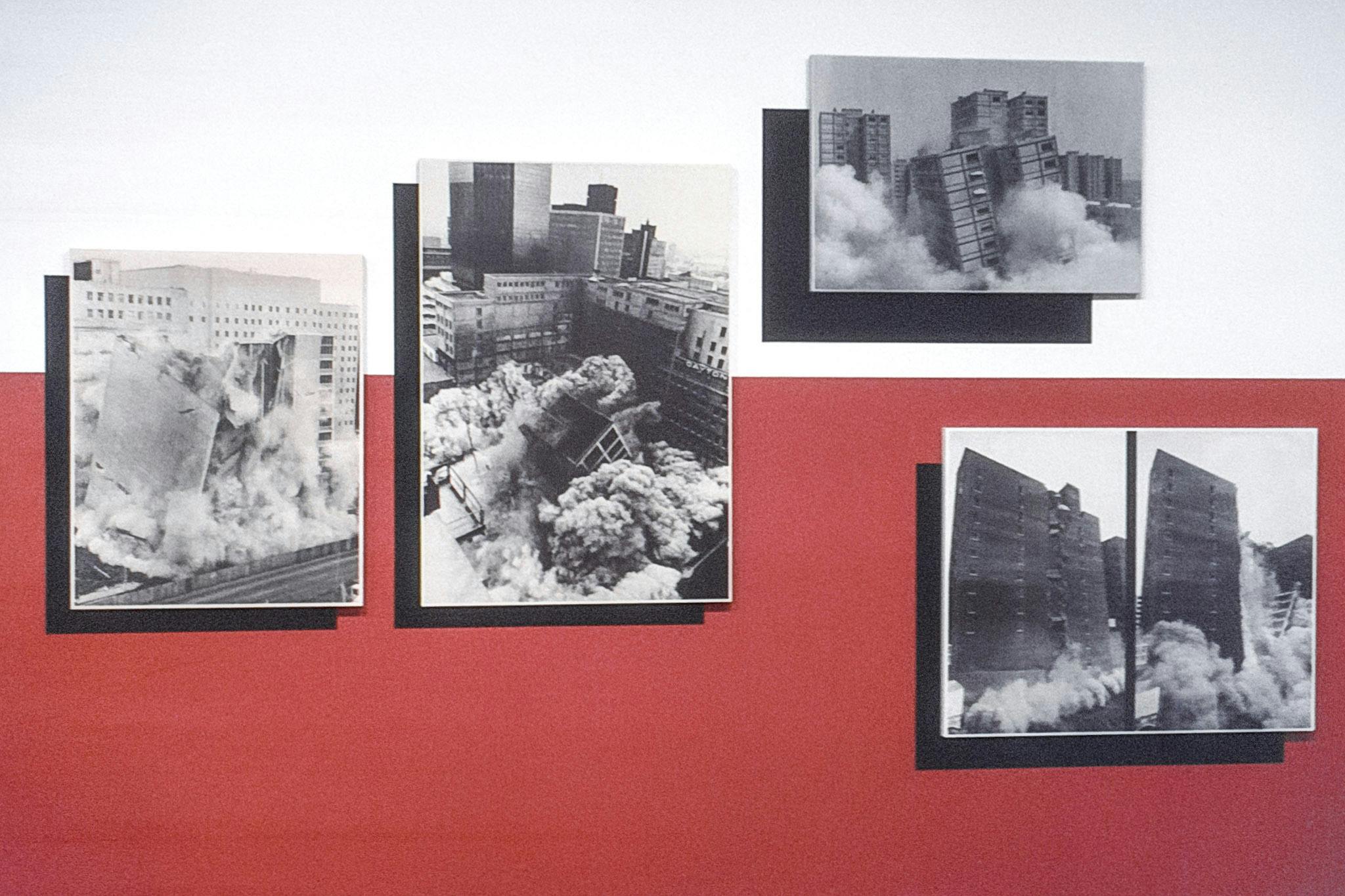A closeup of four black and white photos on a white and red wall. The photos show different large buildings collapsing. The works are mounted slightly away from the wall, creating strong shadows.