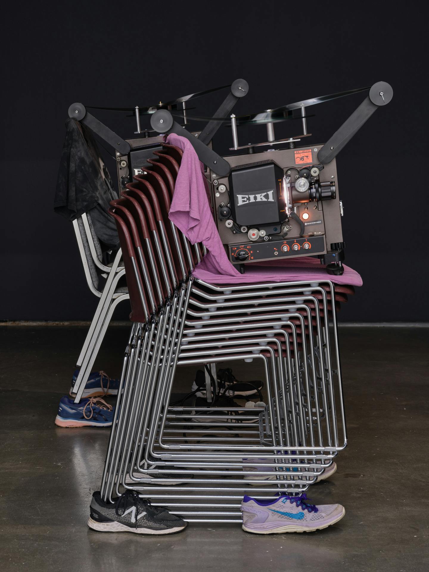A sculpture made of stacked chairs supporting a film projector. A pink t-shirt lines the top chair that the projector sits on, and runnings shoes support the bottom chair.