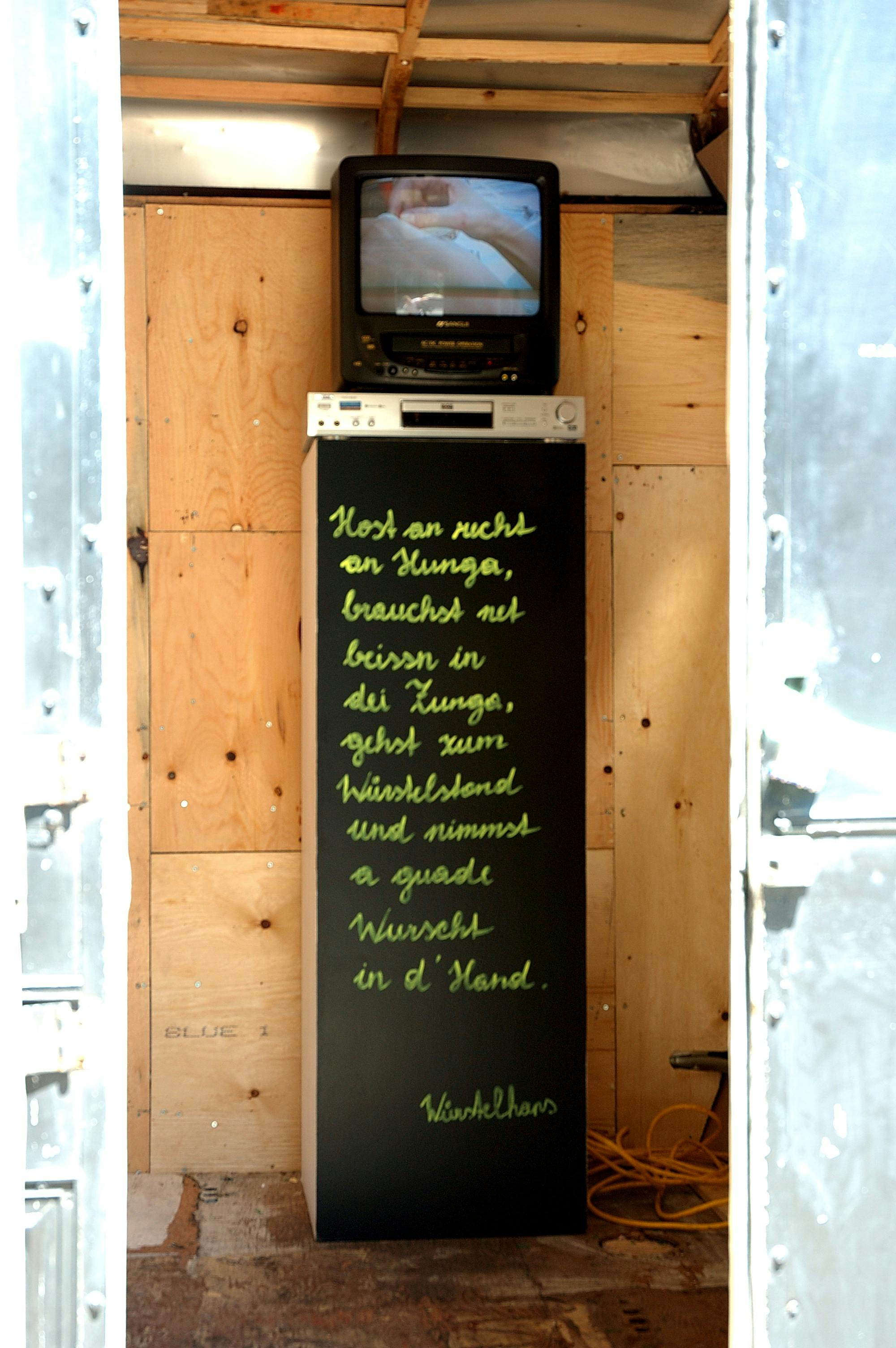 An installation image of an artwork inside storage. A black painted tall pedestal is placed on the floor, on which a poem is handwritten. A CRT TV and a video player are placed on top of this painted pedestal. 