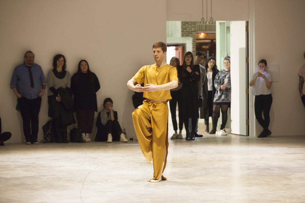 A gallery space with people lined against the wall watching one person perform at the center. The performer is wearing all yellow clothes and is holding a dance pose. 