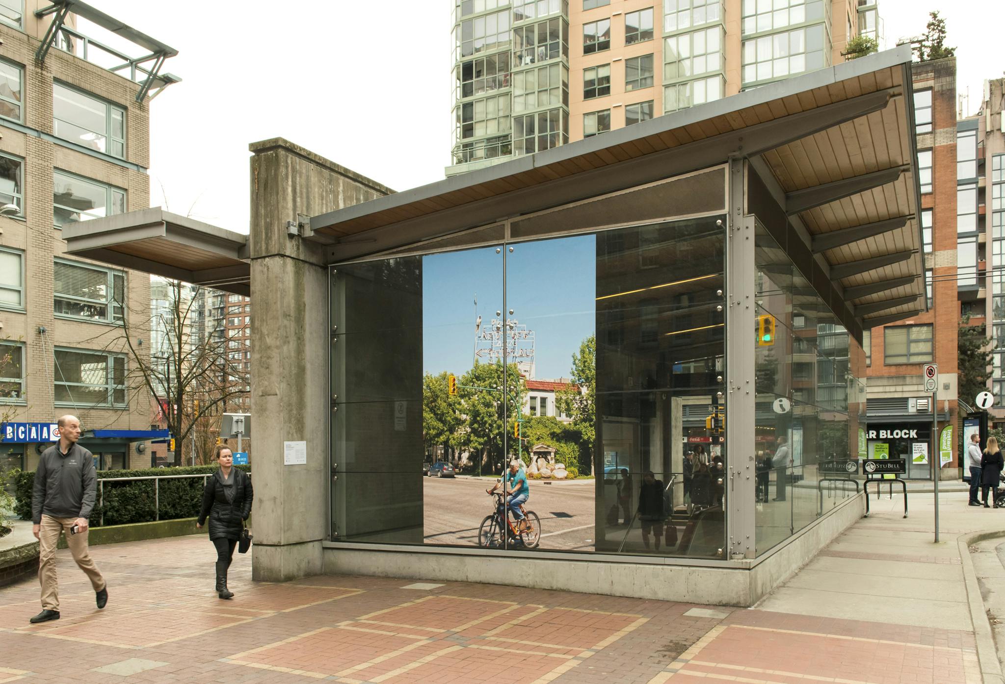 Exterior of the Yaletown Roundhouse Station with a large, vinyl photograph installed on the facade. In the photograph, a person bicycles past a building with a large sign in Cree syllabics on top.