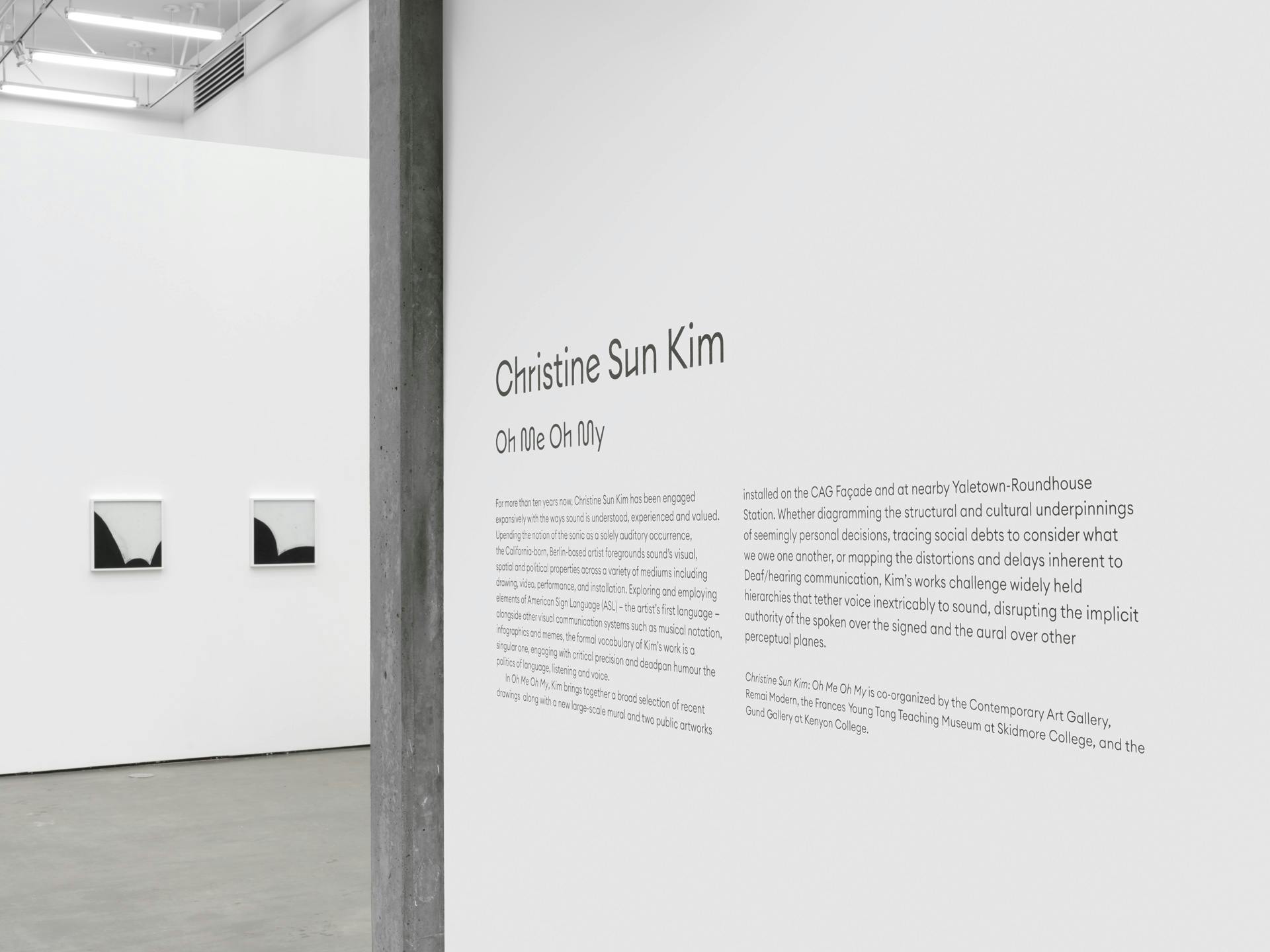 Entryway into a gallery with two drawings by Christine Sun Kim visible on an interior wall. Drawings each feature curved black shapes on a white background.