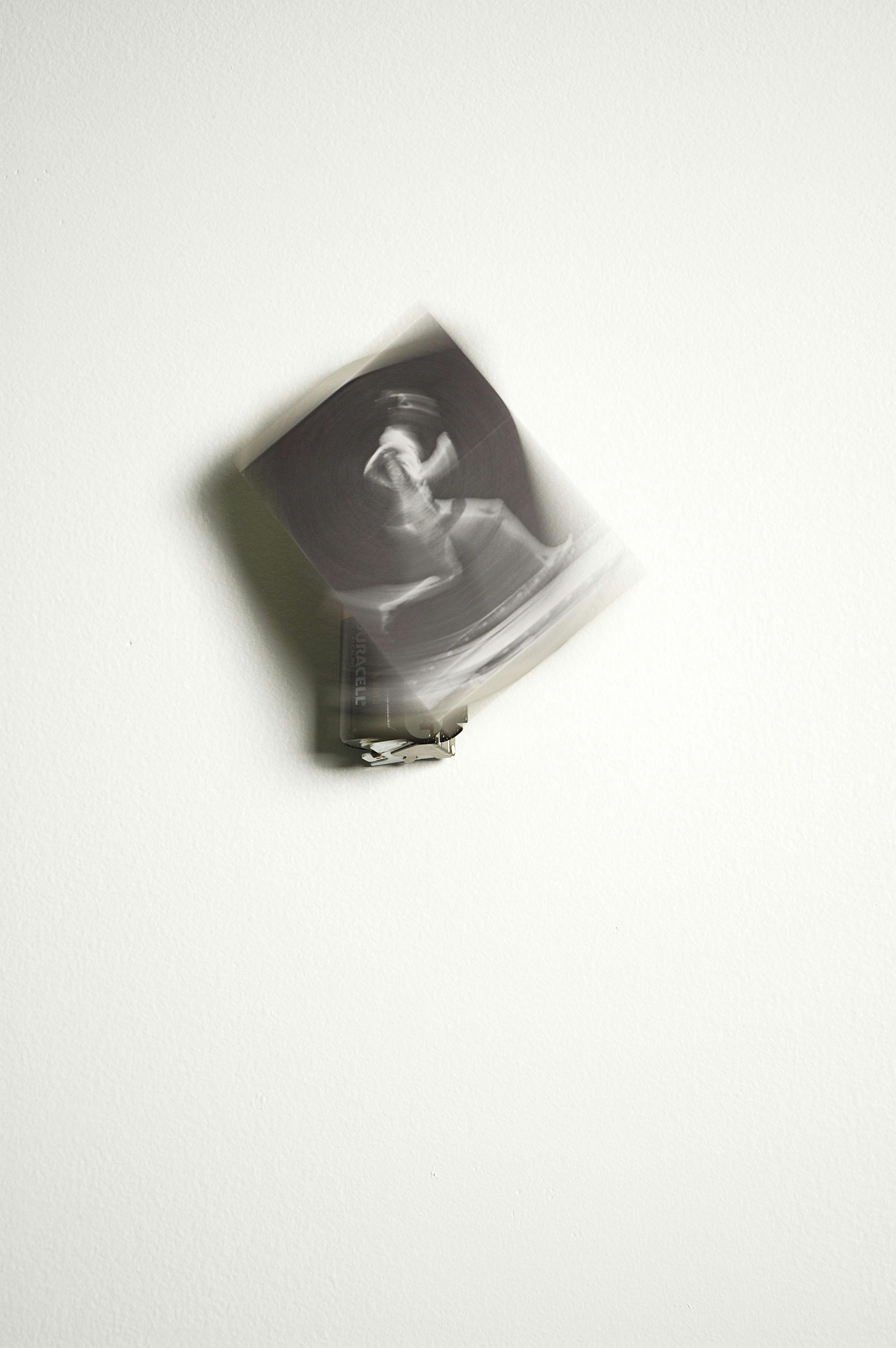 A small black and white photograph of a person running is mounted on a motorized mechanism hanging on a gallery wall. The photograph appears blurred and in motion. 