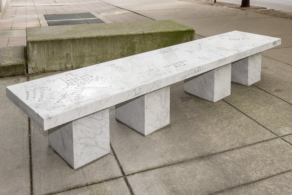 A white marble bench sculpture installed outside the CAG entrance. It is supported by four, thick quadrangular legs. The surface of the long, rectangular seat is covered with carved graffiti. 