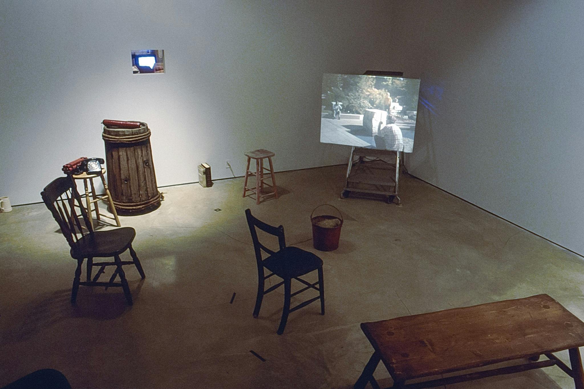 A wooden bench and chairs are placed in the corner of a gallery space. A video piece is projected on a white panel that is placed on a small step stool at the corner of the room.  