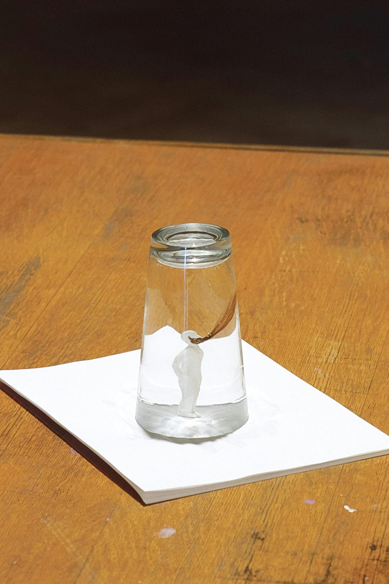 This is a close-up view of an artwork placed on the wooden desk. On a small pile of white paper napkins, a water-filled glass is placed upside down. A small white human figure stands inside the glass.