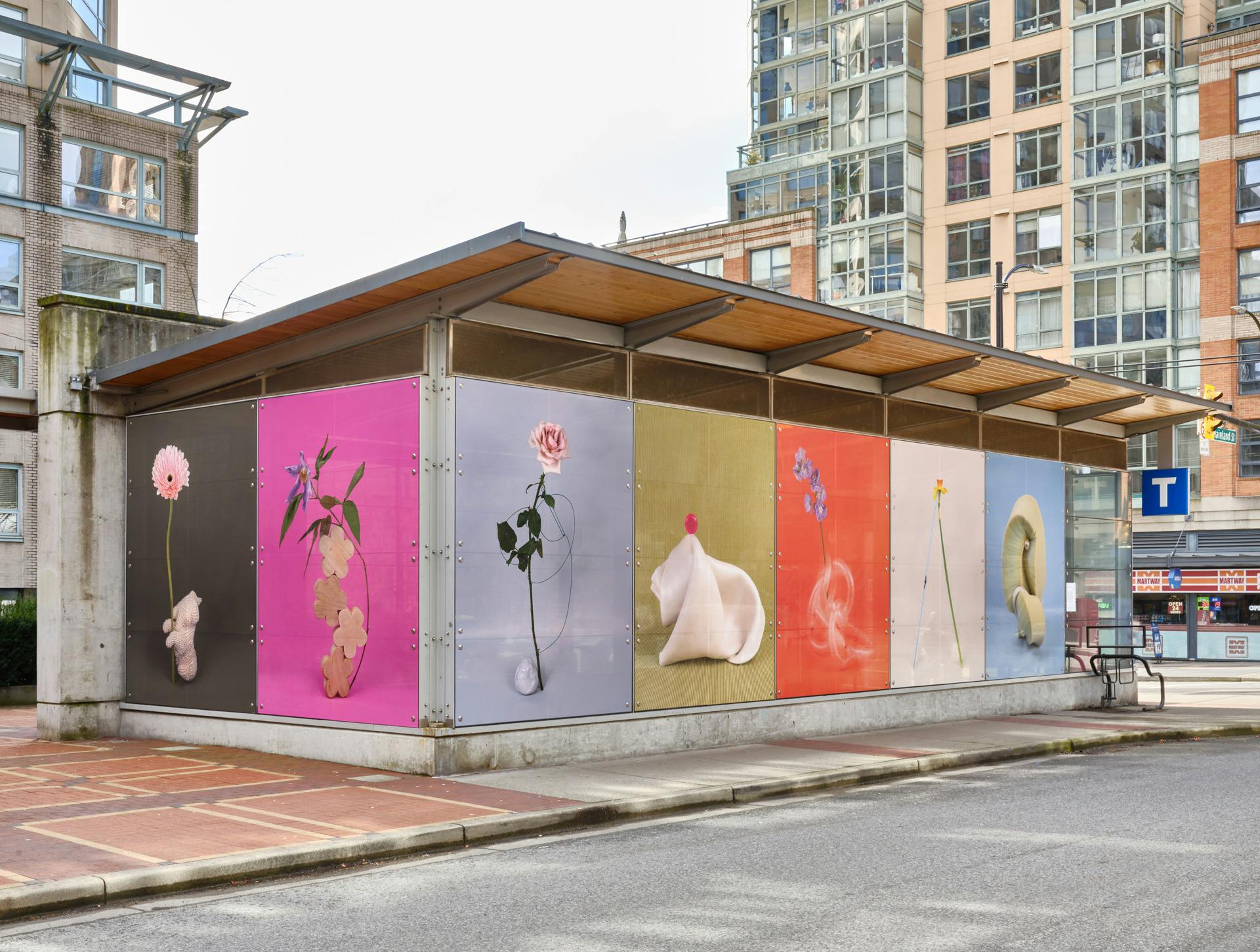A corner view of Yaletown-Roundhouse train station covered entirely in seven large-scale, colourful still-life photographs. The images depict a variety of singular flowers with various objects. 