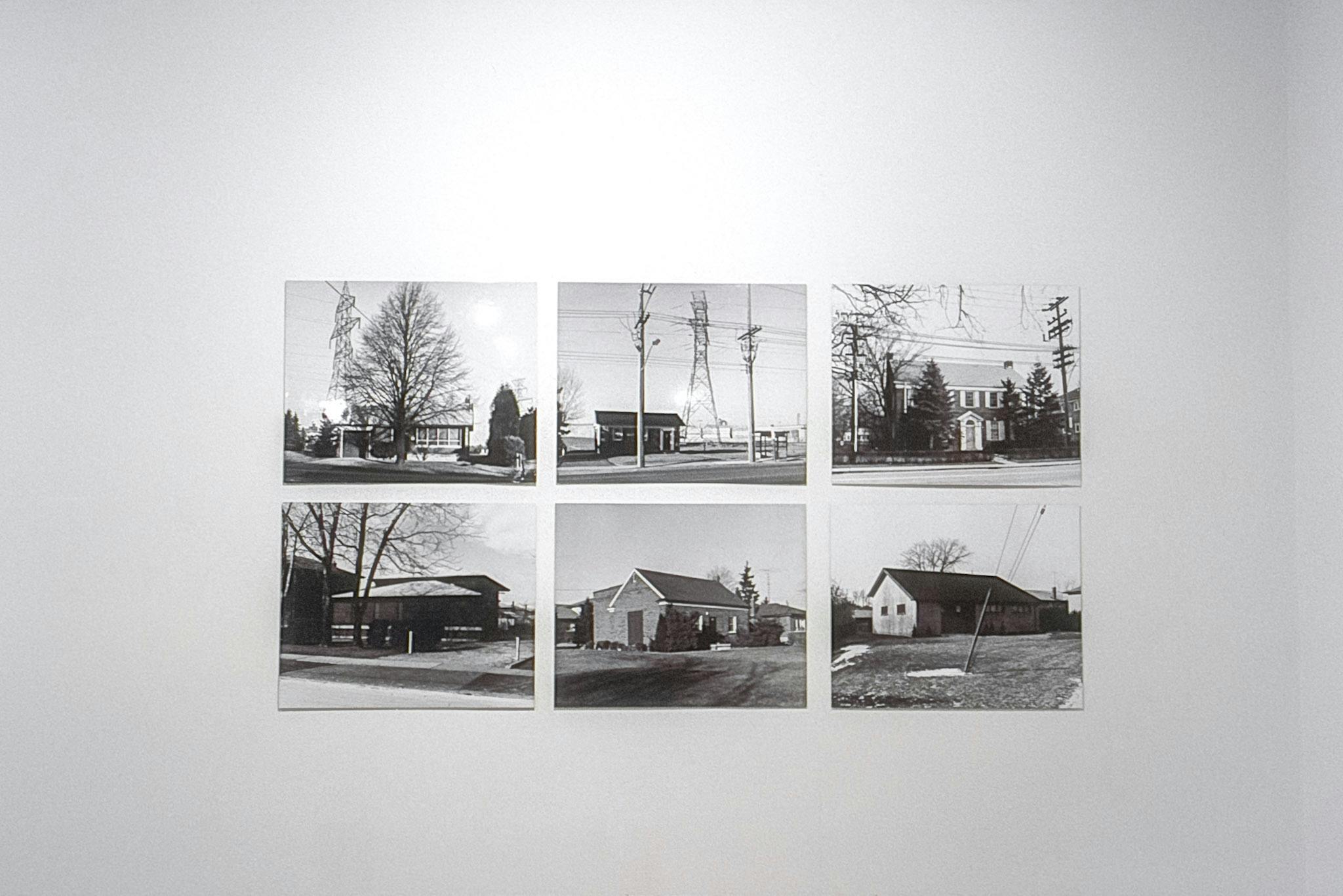 A closeup of 6 unframed black and white photos on a wall. The photos show houses of various sizes in different neighbourhoods. Some have trees in front, and some are in large fields, etc.