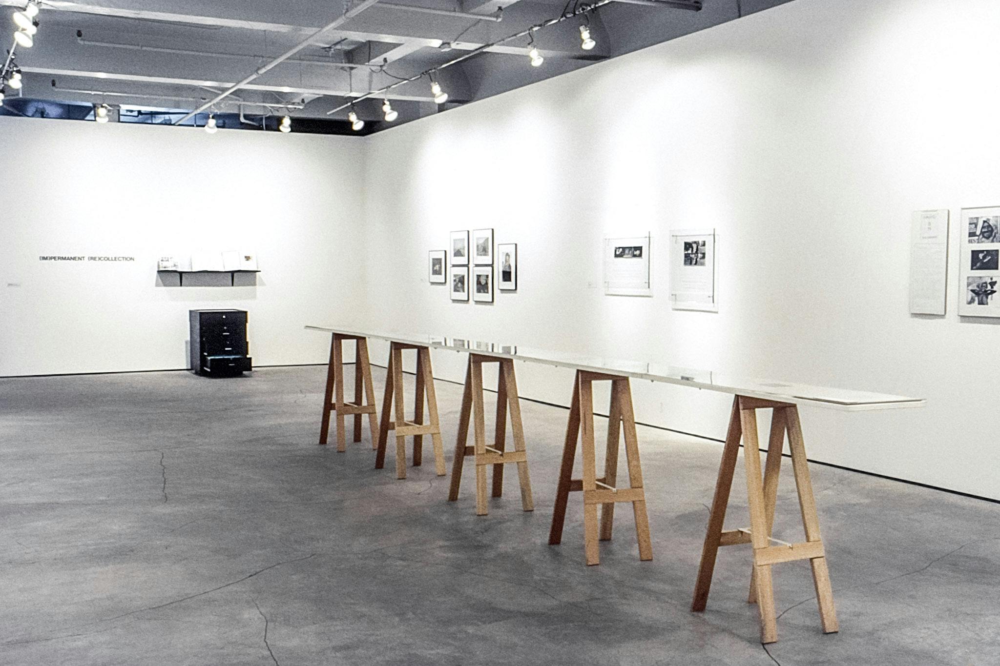 A gallery with framed black and white photos mounted on one wall, and photo prints on textured paper hanging on the other. On the floor, there is a long table-like form with a narrow, glossy top. 