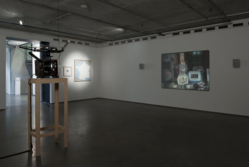 Several artworks installed in a gallery. A film projector projects a moving image of clocks on a large screen mounted on a wall. A map of France is framed and mounted on the other wall.