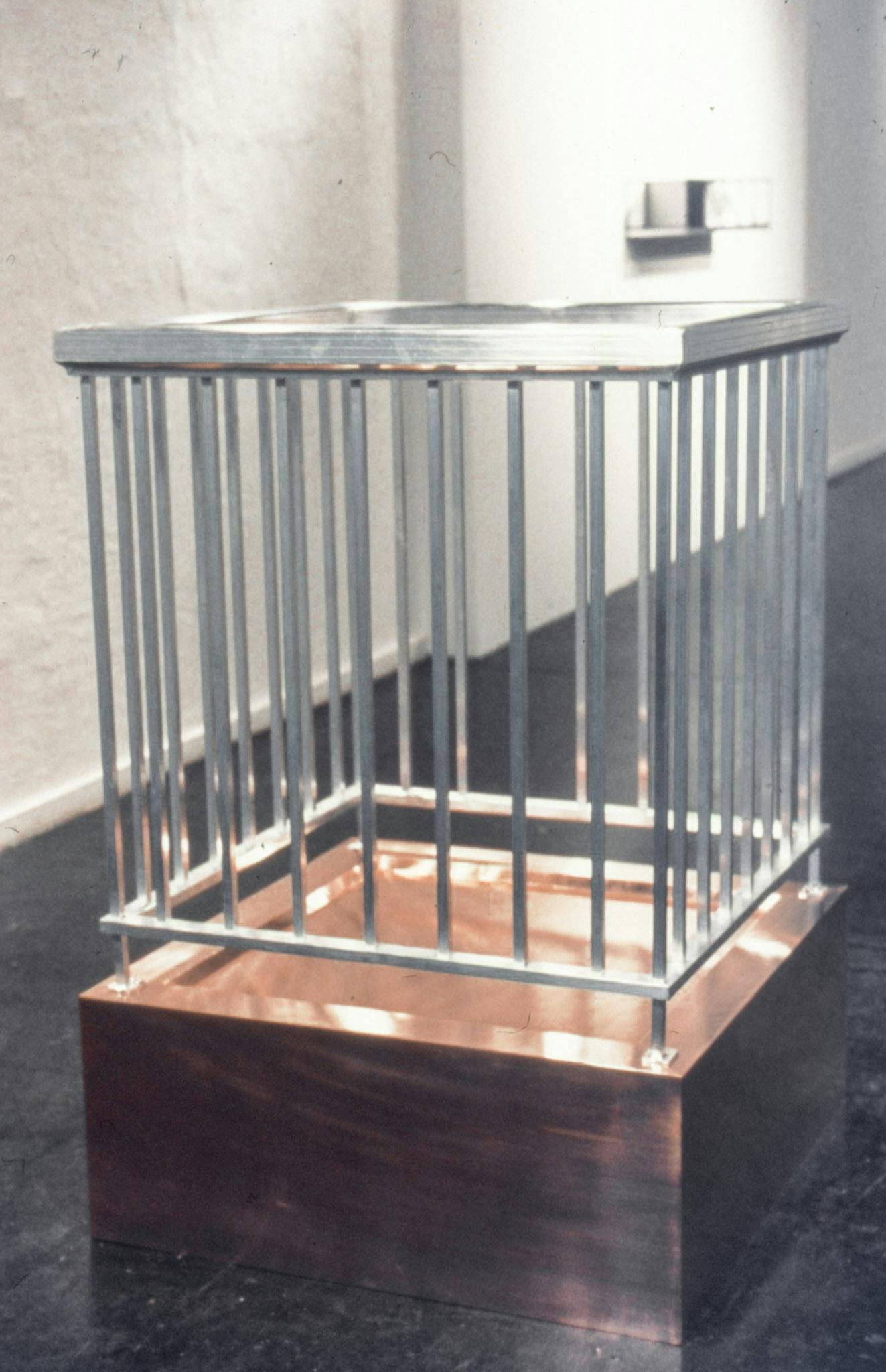 A closeup of an artwork in a gallery. The work is made of a low copper box with a tall silver metal rail attached to the top of the box. Behind it, a work mounted on the white wall is visible.