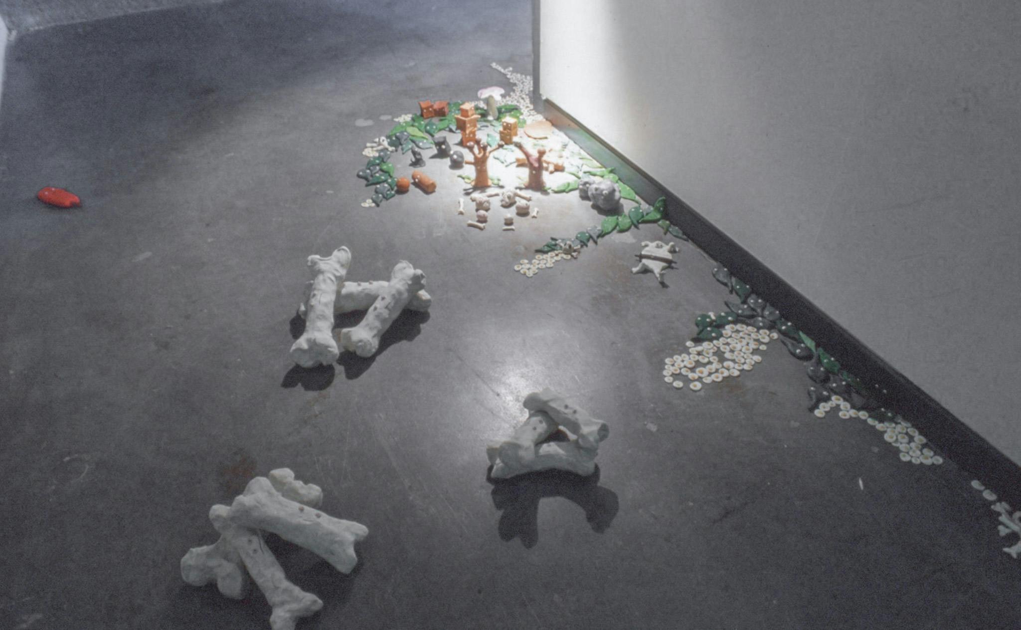 A series of small sculpture-like artworks are installed on a floor. A number of fried-egg shaped objects, bones, and small green round objects are piled up around the corner of the room.