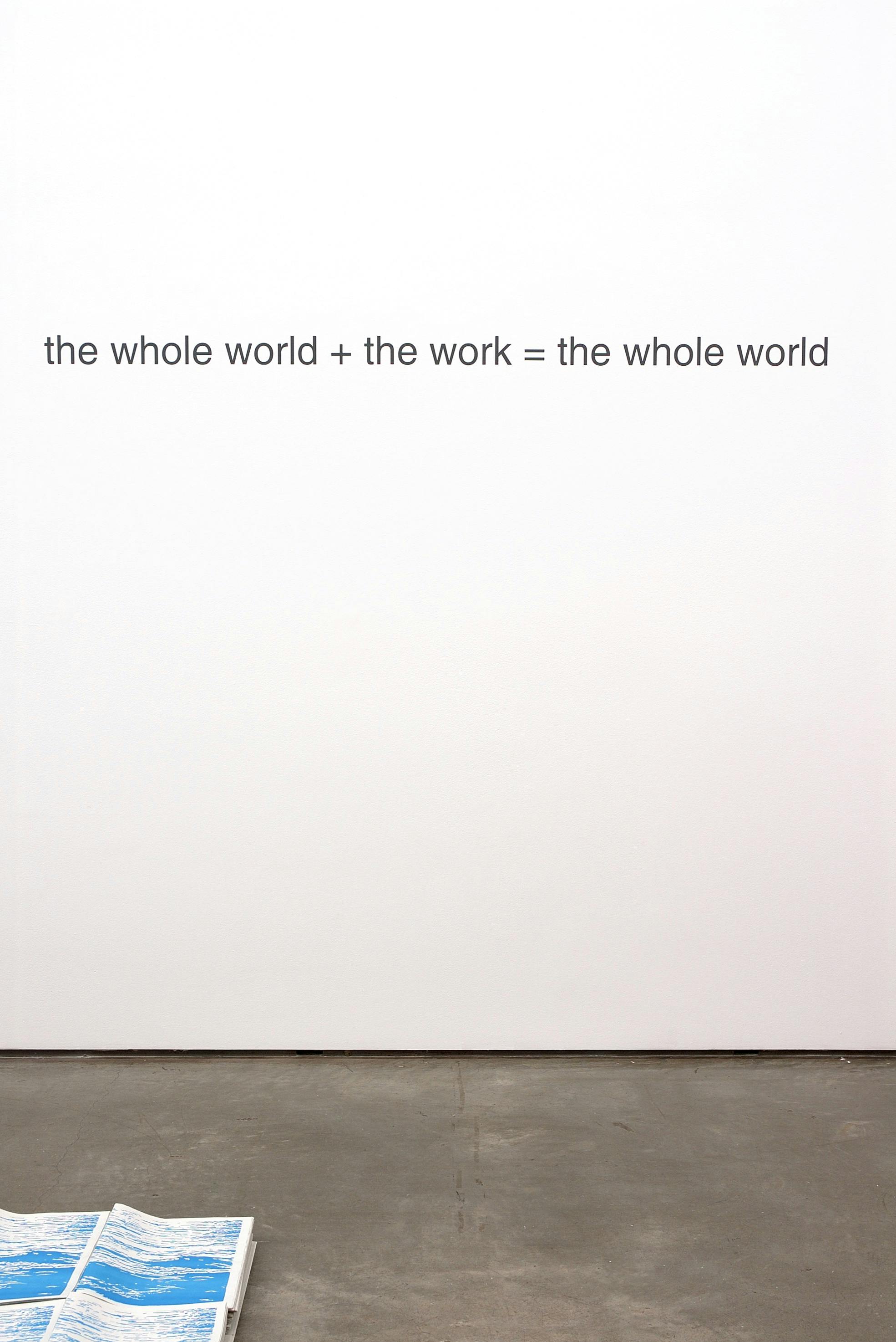A text-based artwork is printed on a gallery wall. It reads, “the whole world + the work = the whole world” in black letters. A collection of blue and white paper is placed on the floor near the sign.