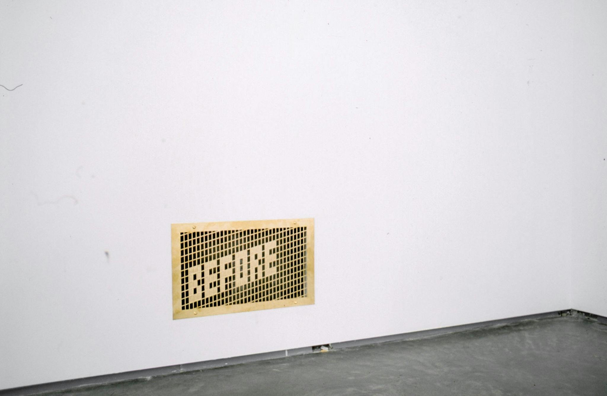 A small piece of brown paper is attached to the bottom of a white wall in a gallery. The image of a word before in capital letters emerging from a black background is printed on this paper.  